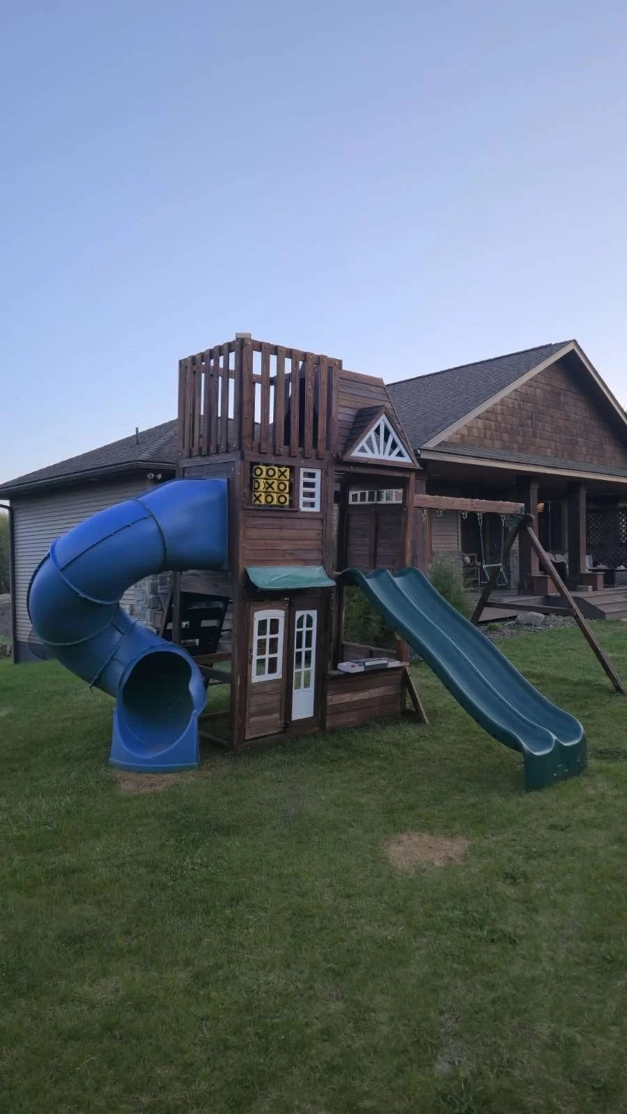 A wooden backyard playset with a blue spiral slide, a green straight slide, a small house, and a deck with a railing, situated on a grassy lawn in front of a house.
