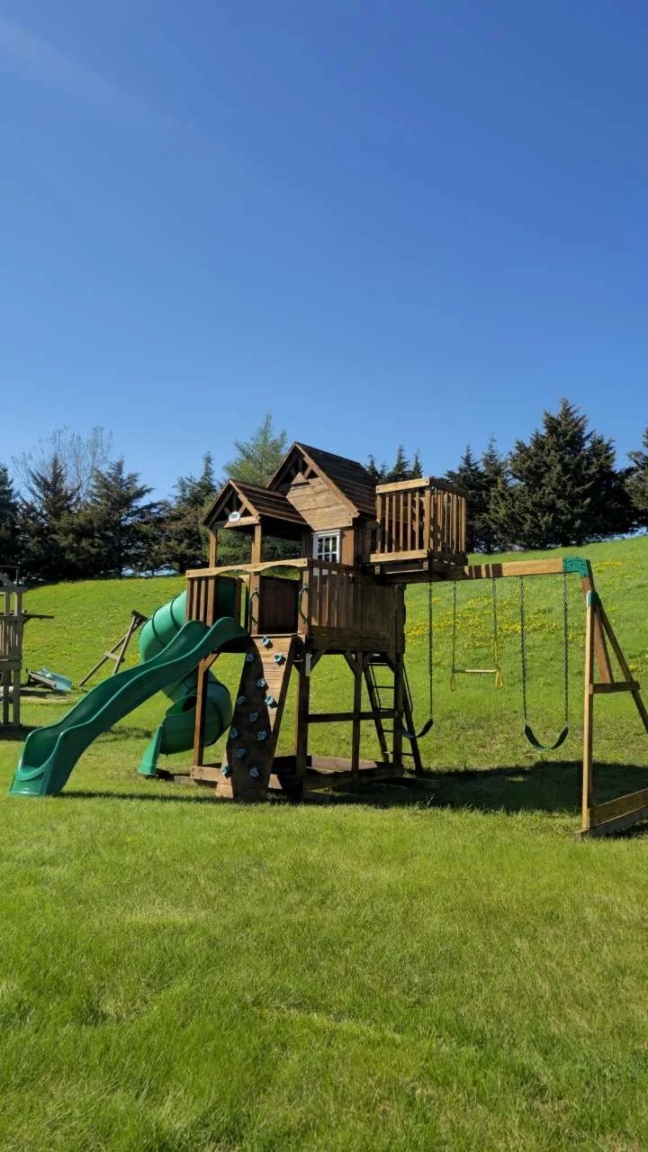 A wooden outdoor playset on a grassy lawn, featuring a green slide, swings, a climbing wall, and a small house with a roof.