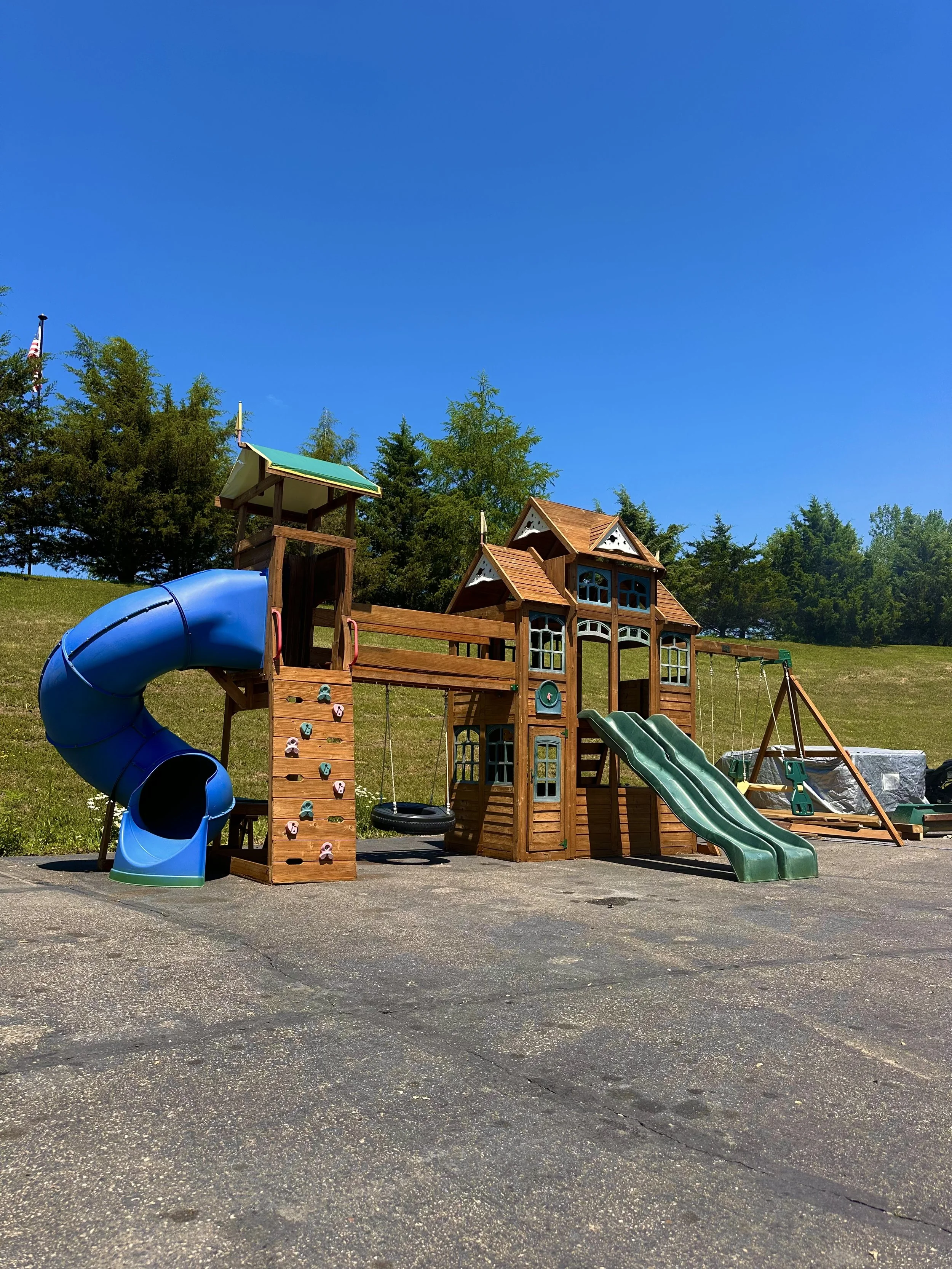 A colorful playground structure with slides, swings, and climbing elements, set on a paved surface with a grassy hill and trees in the background under a clear blue sky.