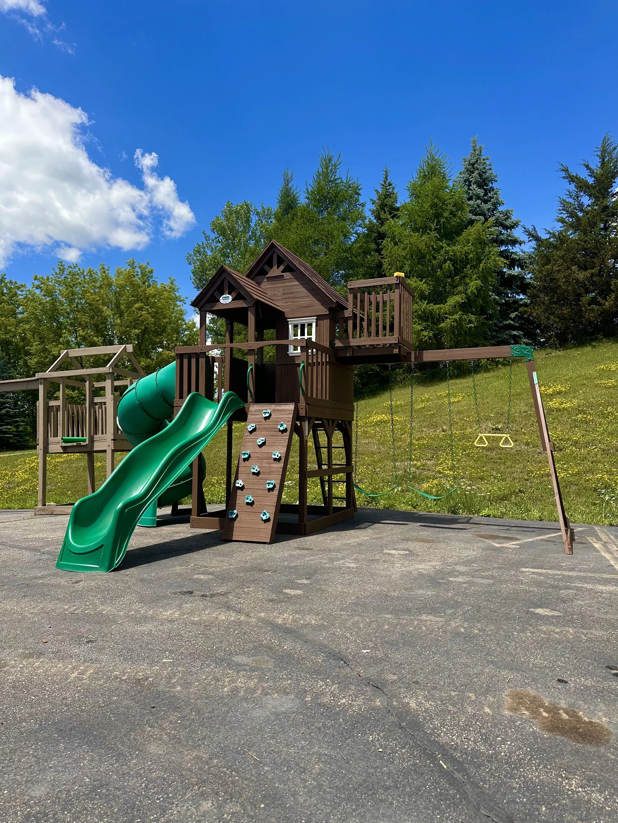 A playground with a wooden playhouse, slides, climbing wall, and swings set on black asphalt, surrounded by green trees under a bright blue sky with white clouds.