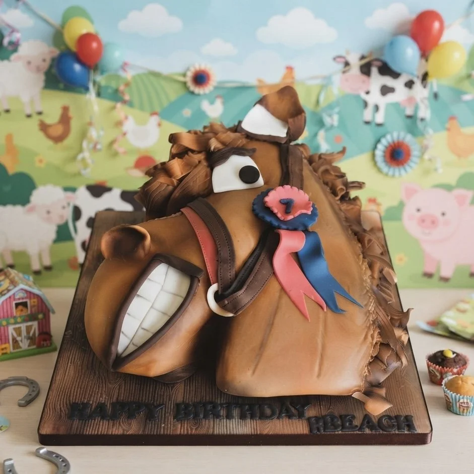Cake shaped like a horse with a brown mane, expressive eyes, a wide toothy grin, and a ribbon. Decorations include farm-themed background with cows, pigs, chickens, and balloons.