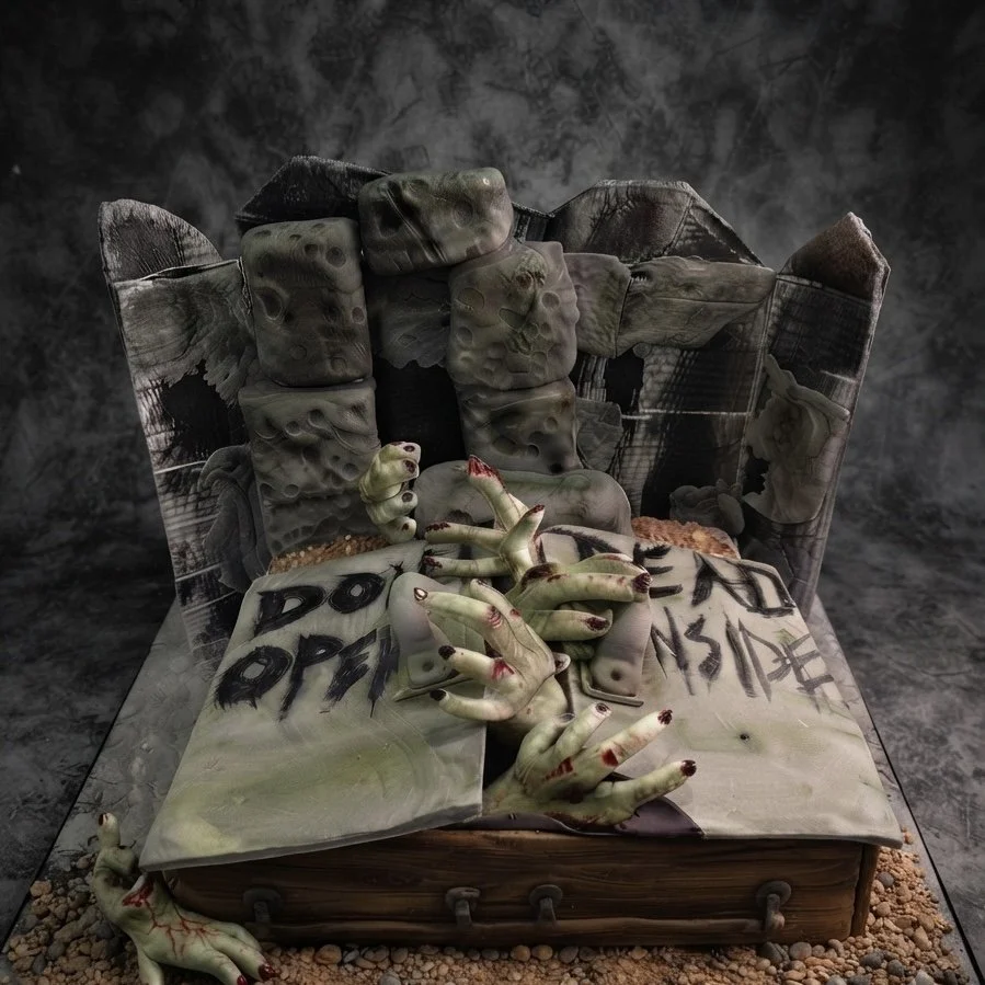 A horror-themed diorama of an open book with blood-stained, zombie-like hands emerging from its pages, set on a wooden base with scattered gravel, featuring a simulated haunted house in the background.