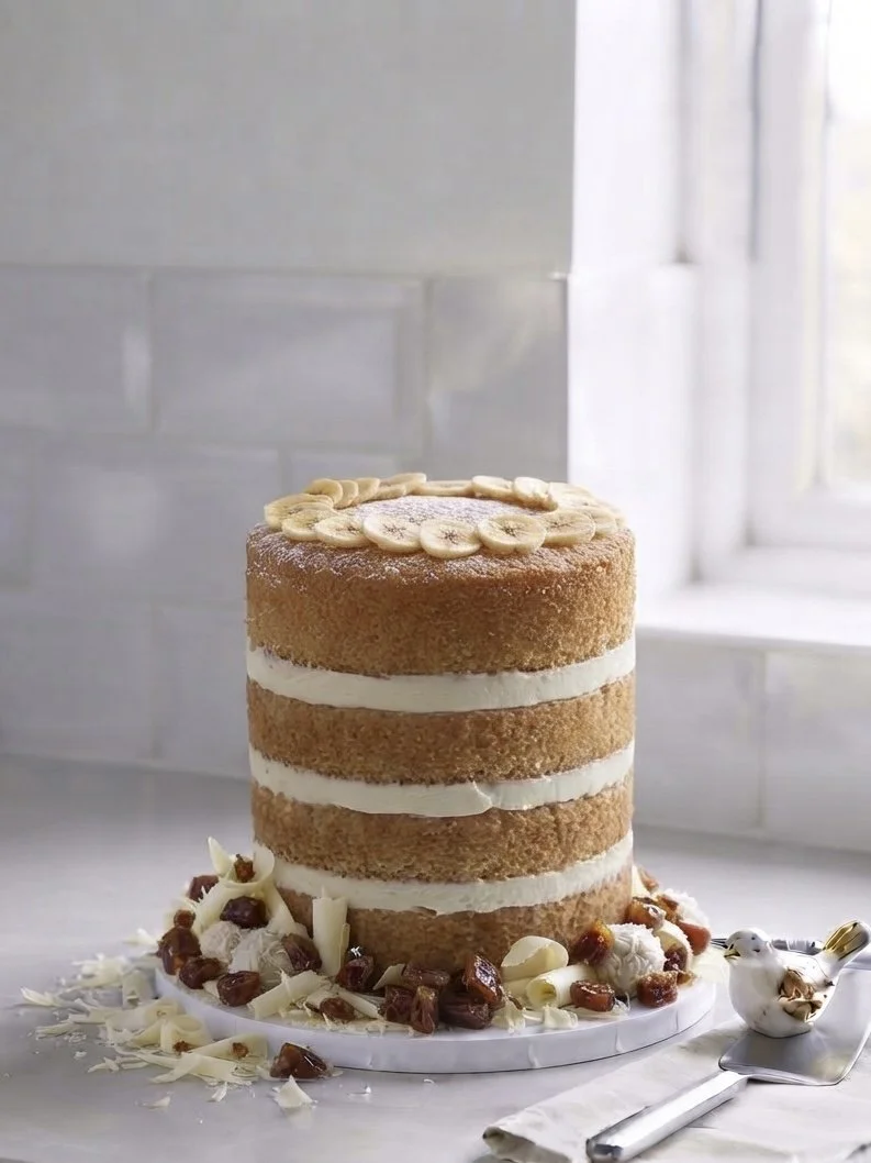 Layered naked cake with banana slices on top, surrounded by white and dark chocolate shavings, dates, and white chocolate curls, set on a marble cake stand near a window.
