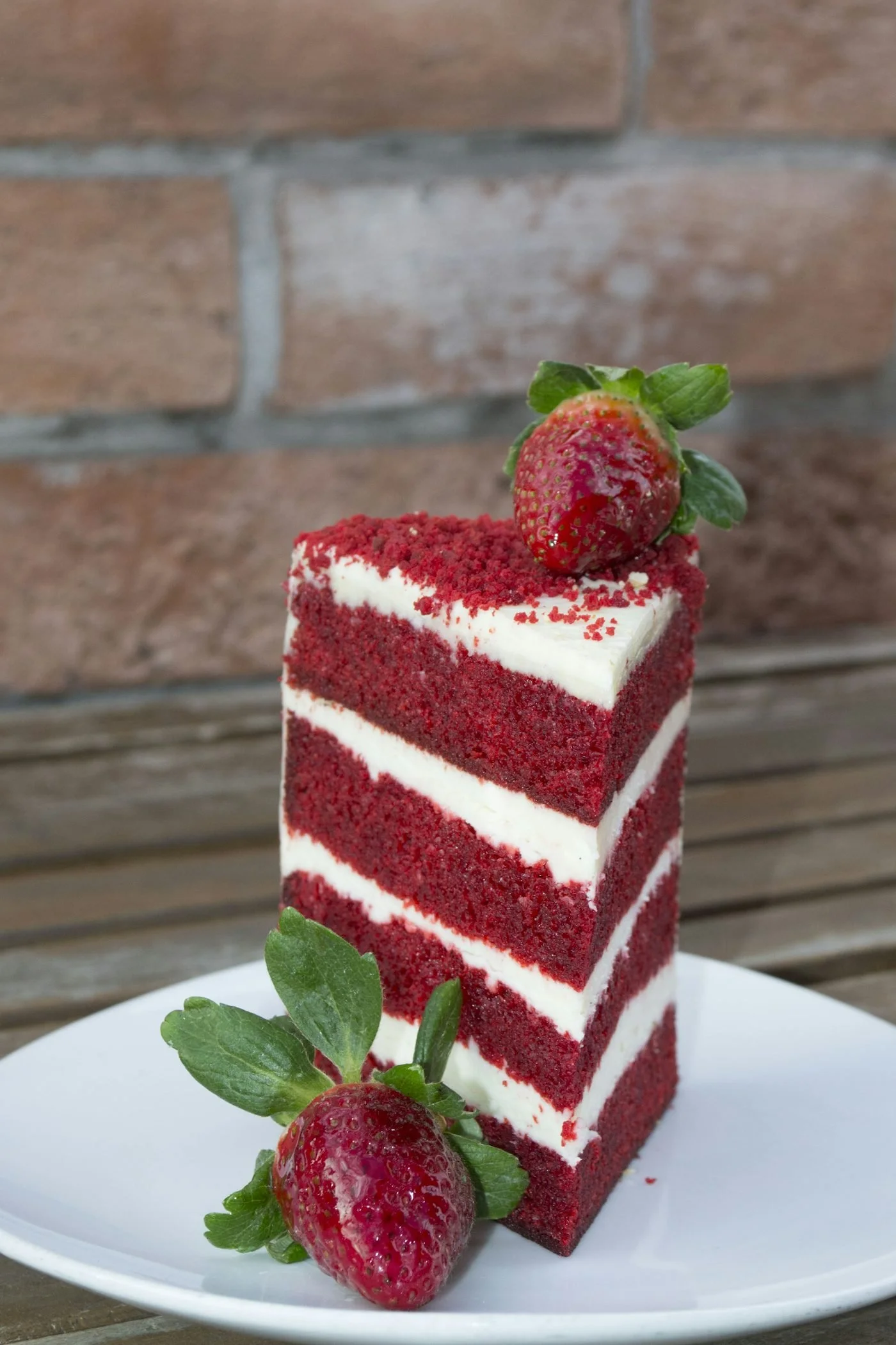 A slice of layered red velvet cake with cream cheese frosting and garnished with strawberries and mint leaves on a white plate