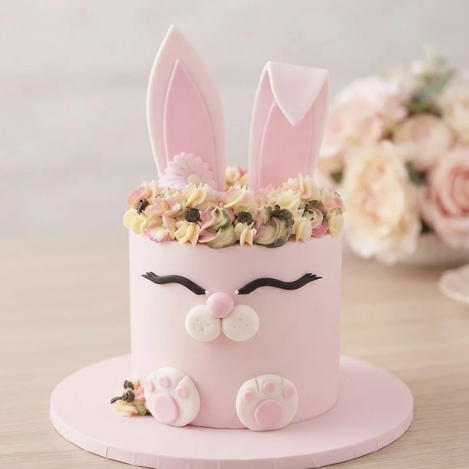 A pink cake decorated to look like a cute bunny with closed eyes, a pink nose, white cheeks, and paw prints, topped with pink and white bunny ears and surrounded by pastel-colored flower-like frosting with small chocolate accents.