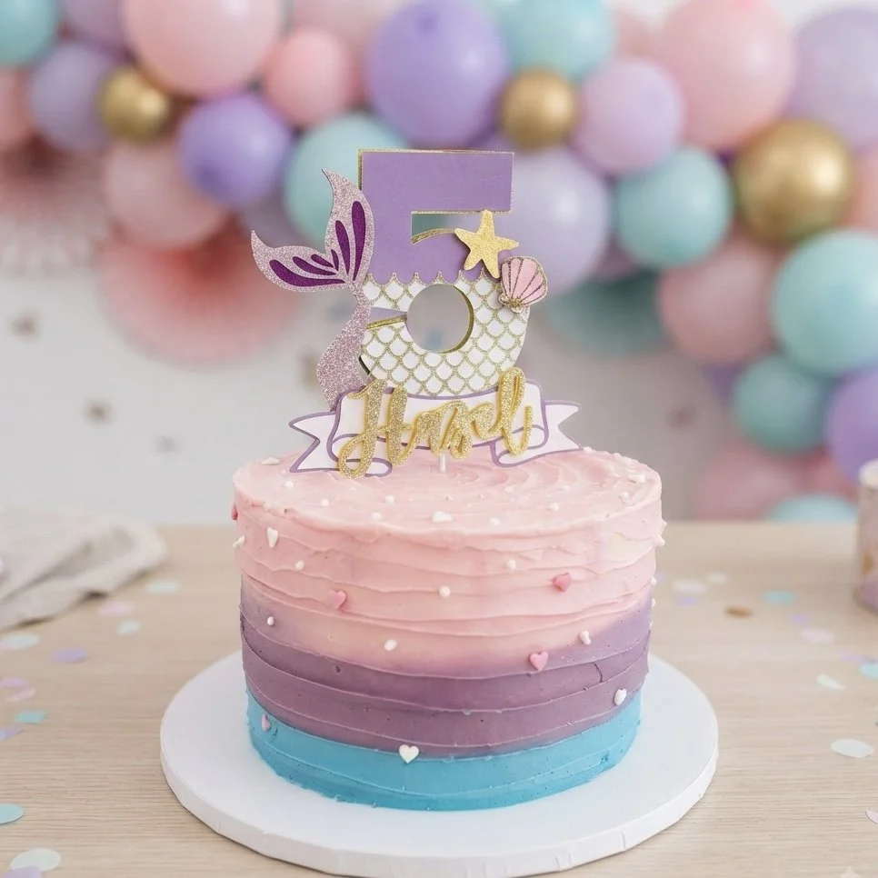 A colorful birthday cake with pink, purple, and blue frosting layers, topped with a decorative unicorn and the number five, surrounded by pastel balloon decorations in the background.