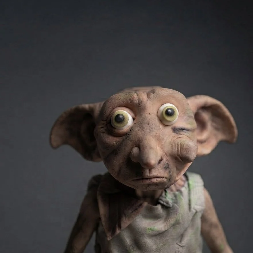 A creature with large ears, big googly eyes, and a wrinkled face, wearing a dirty shirt, against a dark background.