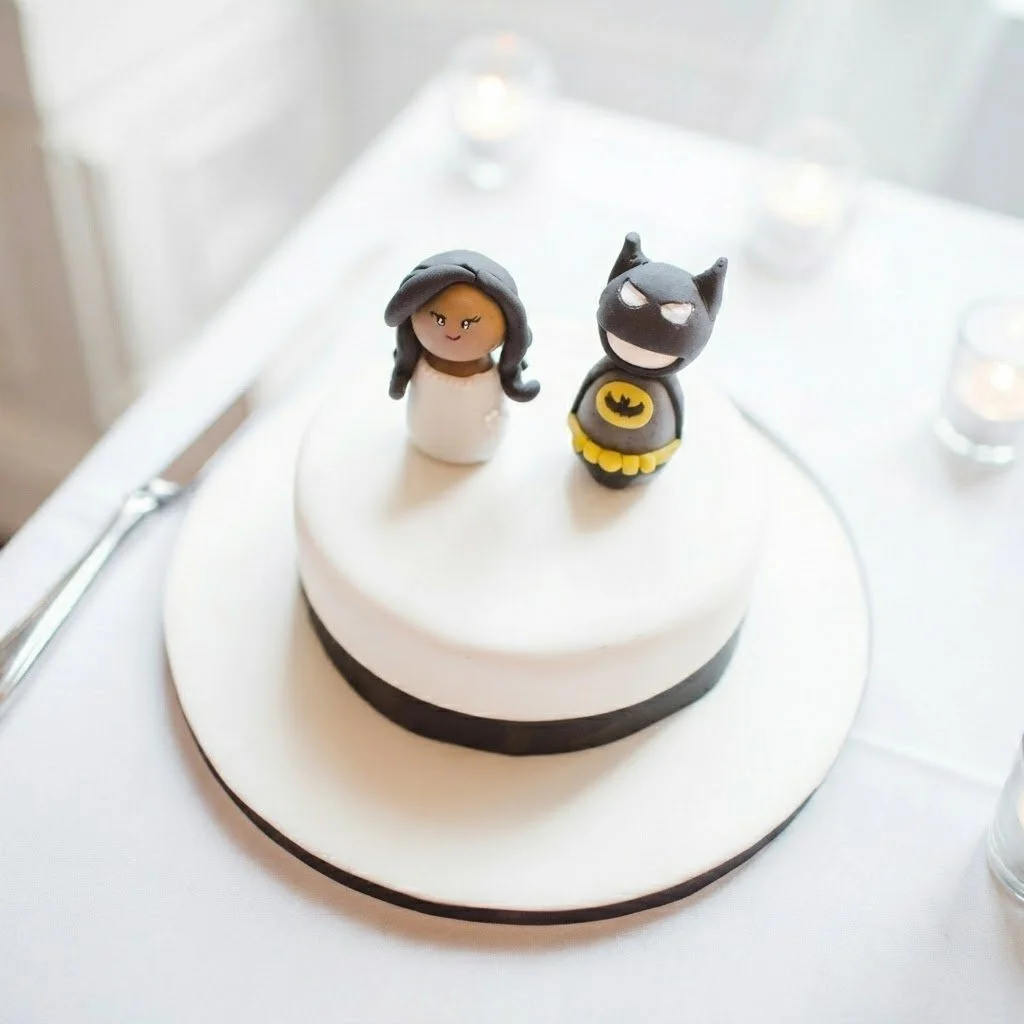 Bespoke white and black Batman themed celebration cake topped with girl and cat figurines. Handcrafted custom cakes designed for unique special occasions.