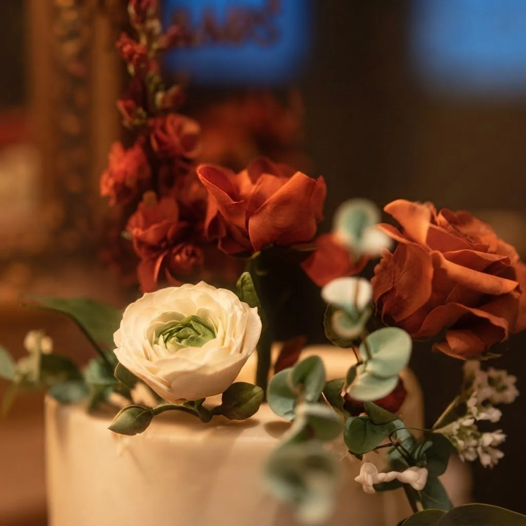 Elegant floral display featuring a white ranunculus alongside deep brownish-red roses and foliage. Handcrafted luxury floral styling for bespoke events.