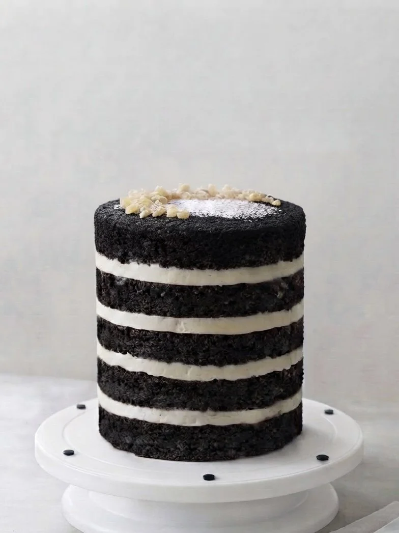 A tall, layered black and white striped cake on a white cake stand, decorated with white chocolate chips on top.