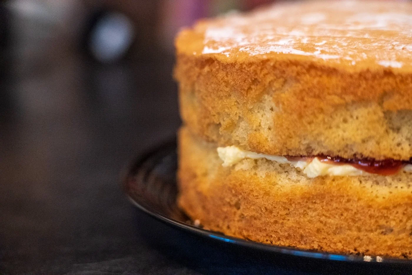 Close-up of a slice of cake with light brown sponge layers and white cream filling, dusted with powdered sugar, on a dark plate.