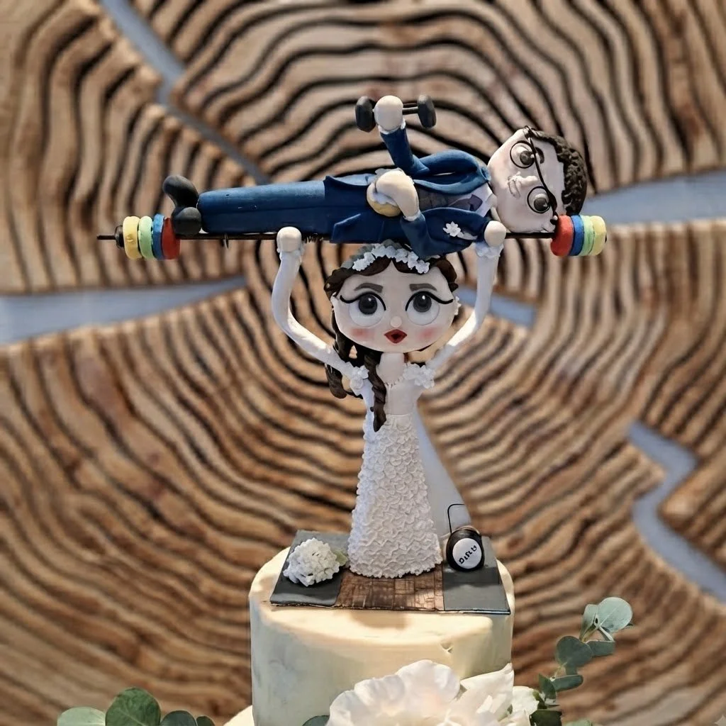 Playful cartoon bride carrying groom cake topper set against a rustic wooden backdrop. Handcrafted bespoke cake toppers designed for unique wedding celebrations.