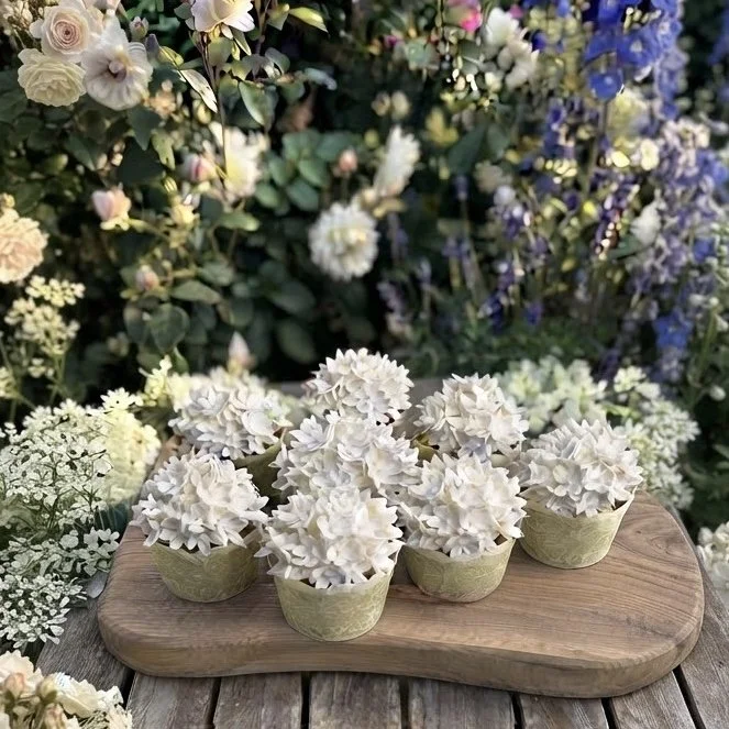 Delicate white flower-shaped desserts served in small green cups on a rustic wooden tray. Handcrafted luxury treats designed for bespoke celebrations.
