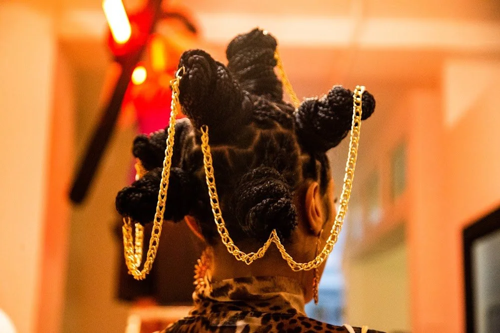 Back of a person with intricately styled hair wrapped in large twists, decorated with gold chains, in an indoor setting.