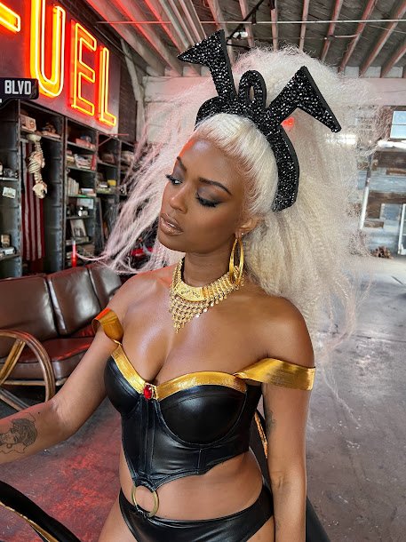 Woman with platinum blonde hair wearing bunny ears headband, black leather strapless top, gold jewelry, and sitting on a couch in a diner with neon signs.