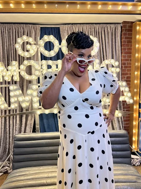 A woman in a white dress with black polka dots, wearing red lipstick and sunglasses, is posing cheerfully in front of a decorated backdrop with the words "Good Vibes" illuminated.