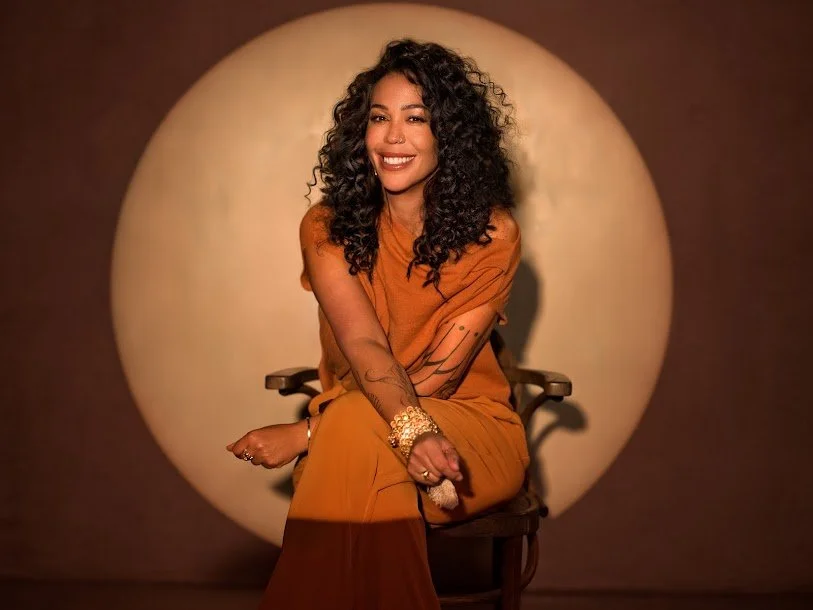 A woman with curly black hair sitting on a chair, smiling, wearing a rust-colored outfit and gold jewelry, with a circular background in a warm tone.