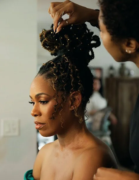 A woman with styled curly hair is having her hair arranged by a hairstylist in a salon or backstage setting.
