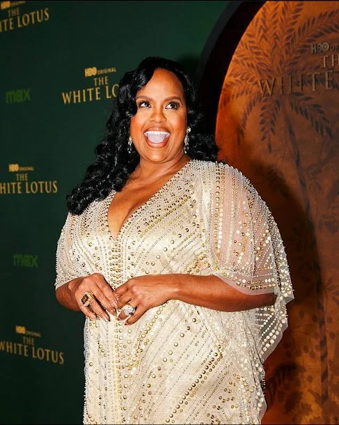 A woman in a sparkling gown posing at an event for 'The White Lotus' with a green backdrop and a wooden arch.