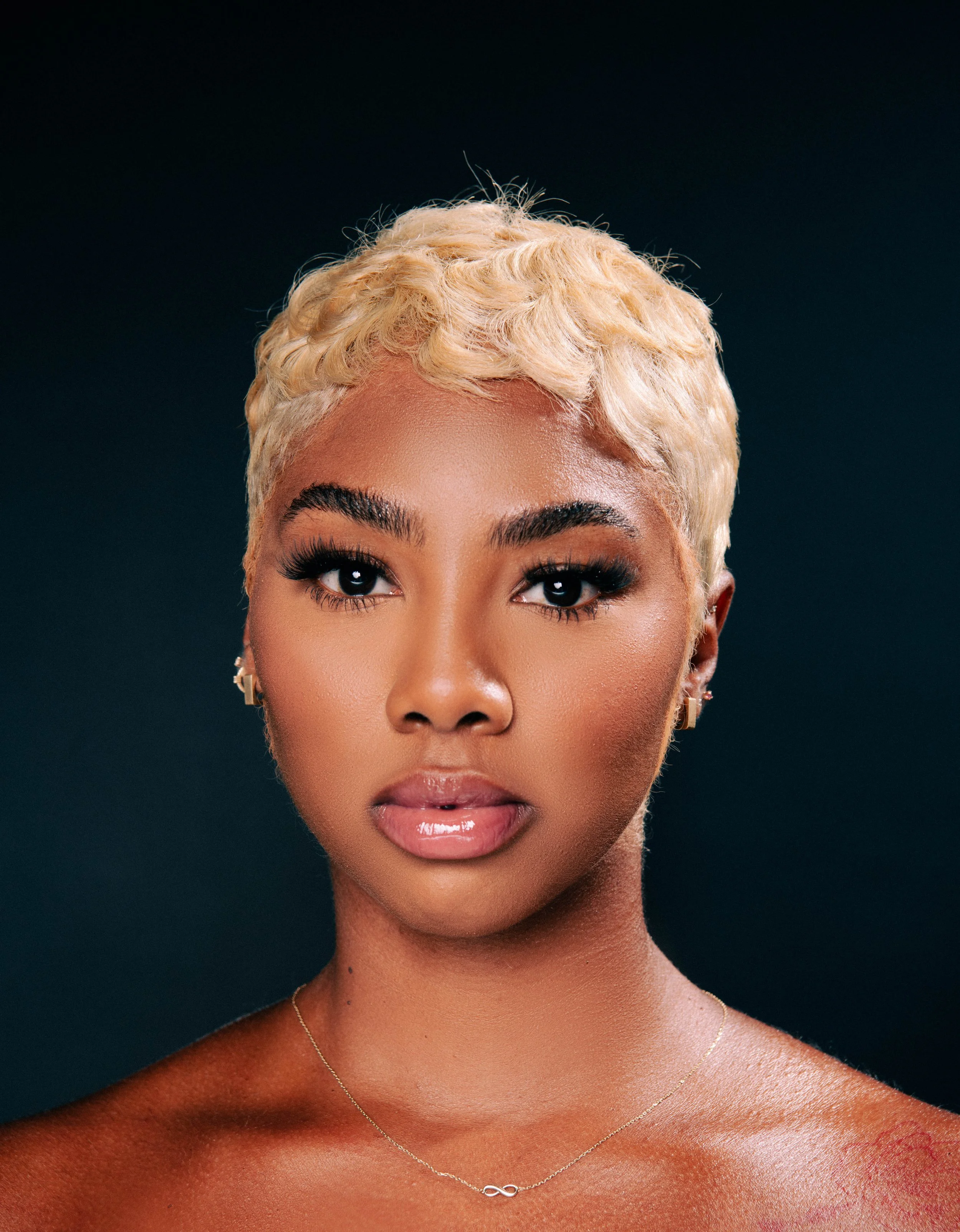 A close-up portrait of a woman with short, curly platinum blonde hair, well-defined eyebrows, and piercing eyes, wearing gold jewelry, set against a dark background.