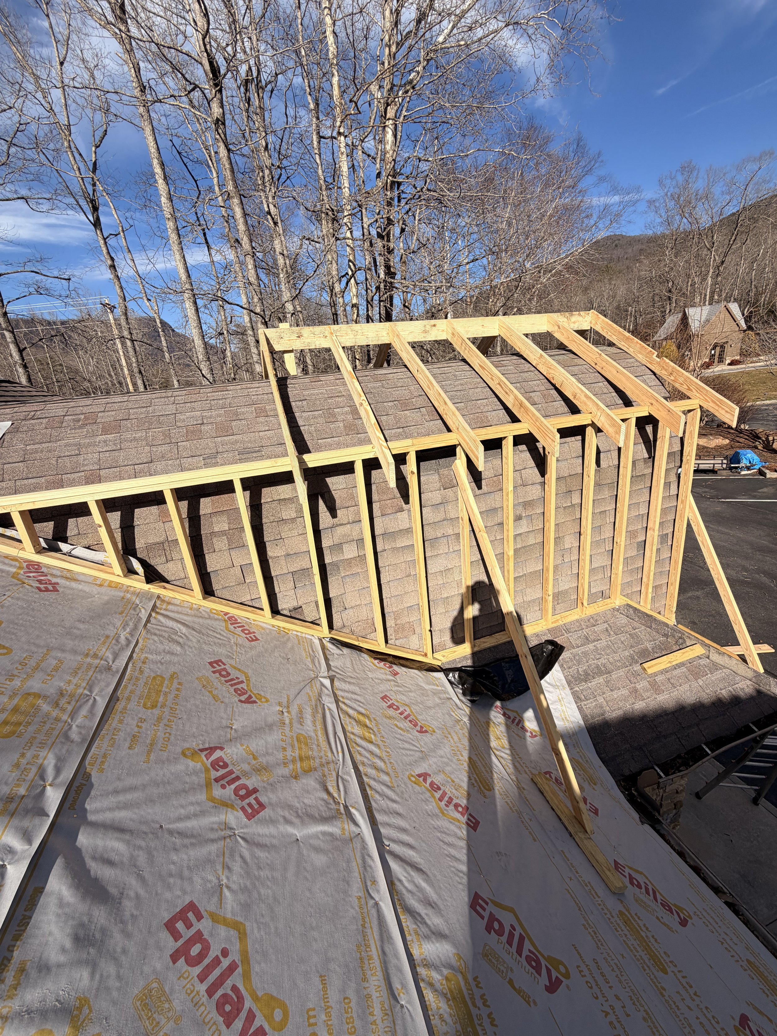 A house roof under construction with visible wooden framing and shingles for the roof. Surrounding trees and a clear blue sky with some clouds in the background.