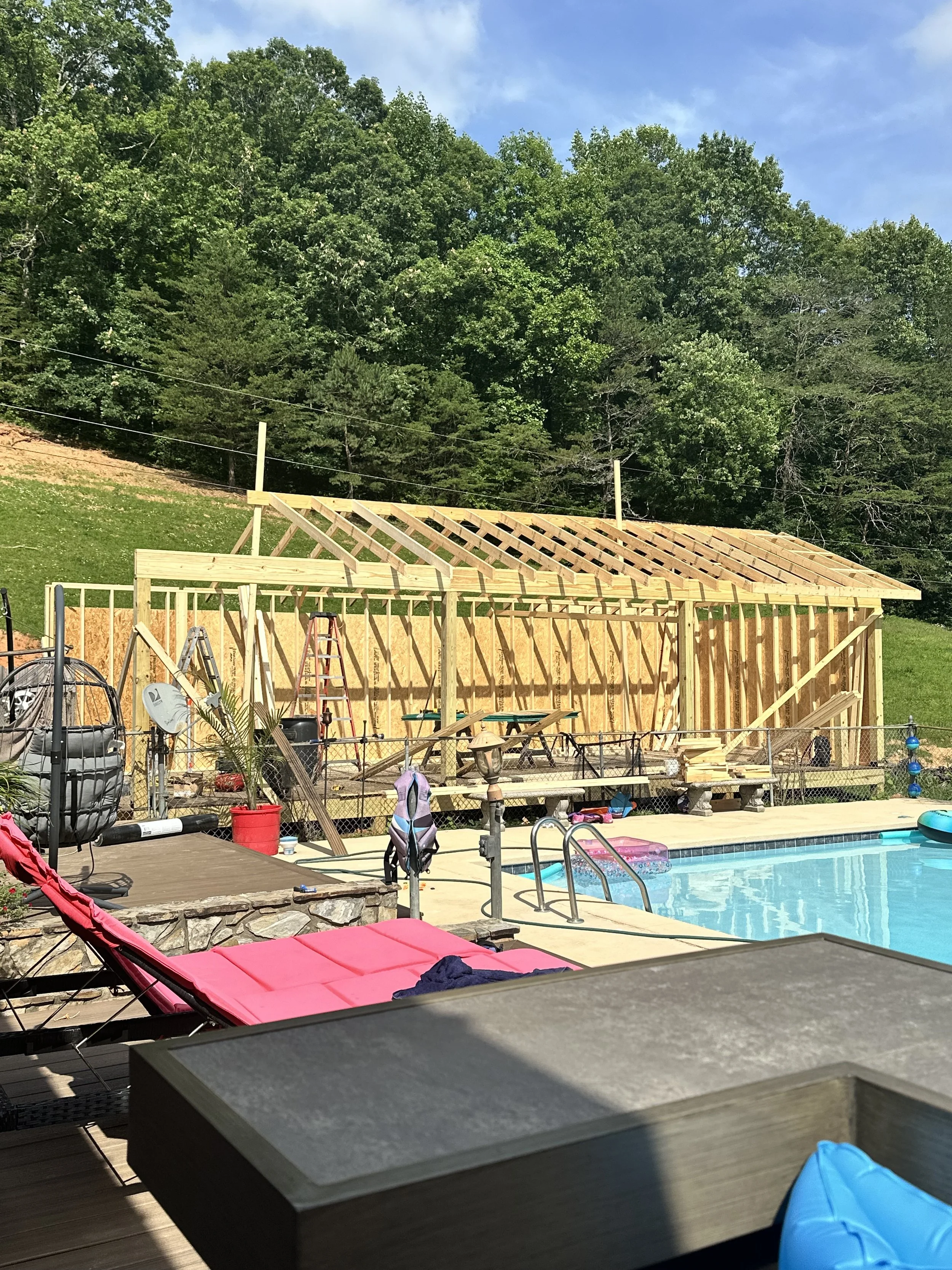 Backyard scene showing a wooden deck under construction, a swimming pool, pool toys, lounge chairs, and a background of lush green trees under a partly cloudy sky.