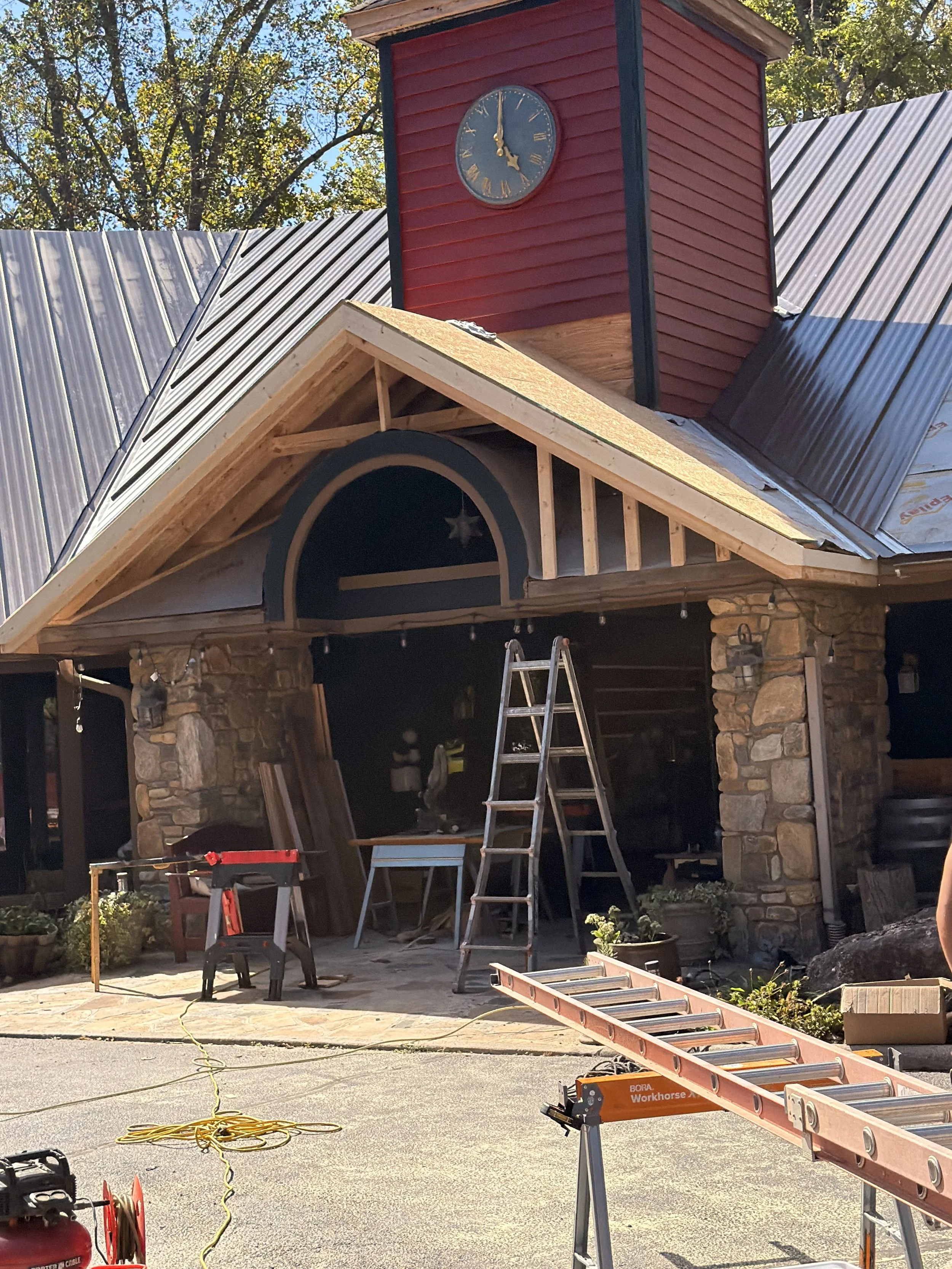 A house under renovation with a stone facade, a partially built gable, and a red clock tower. Construction tools, ladders, and cords are scattered around the yard.