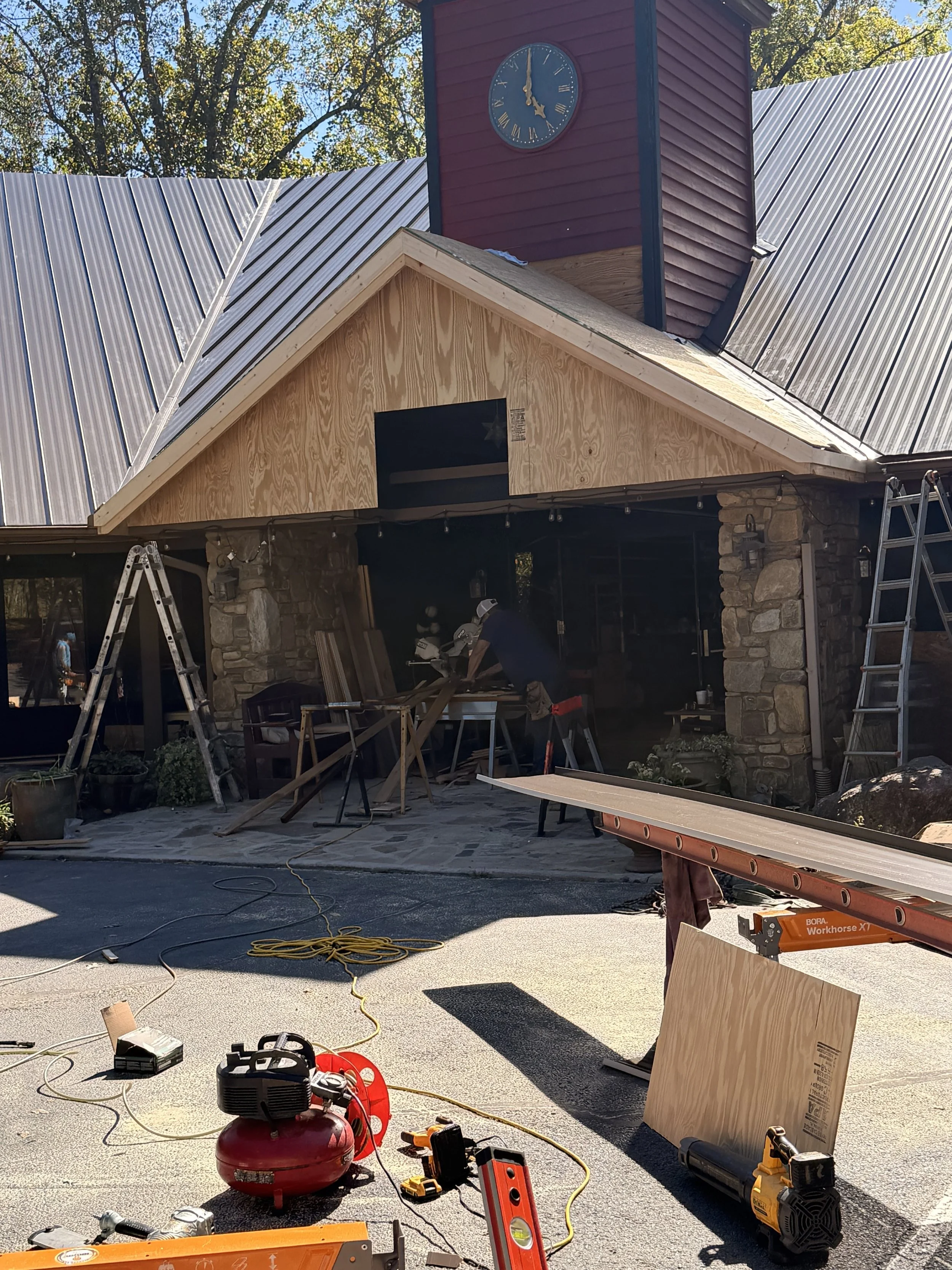 Construction workers are building or renovating a house, with construction tools, ladders, and materials visible in the foreground. The house has a stone chimney, wooden siding, and a changing or unfinished roof.