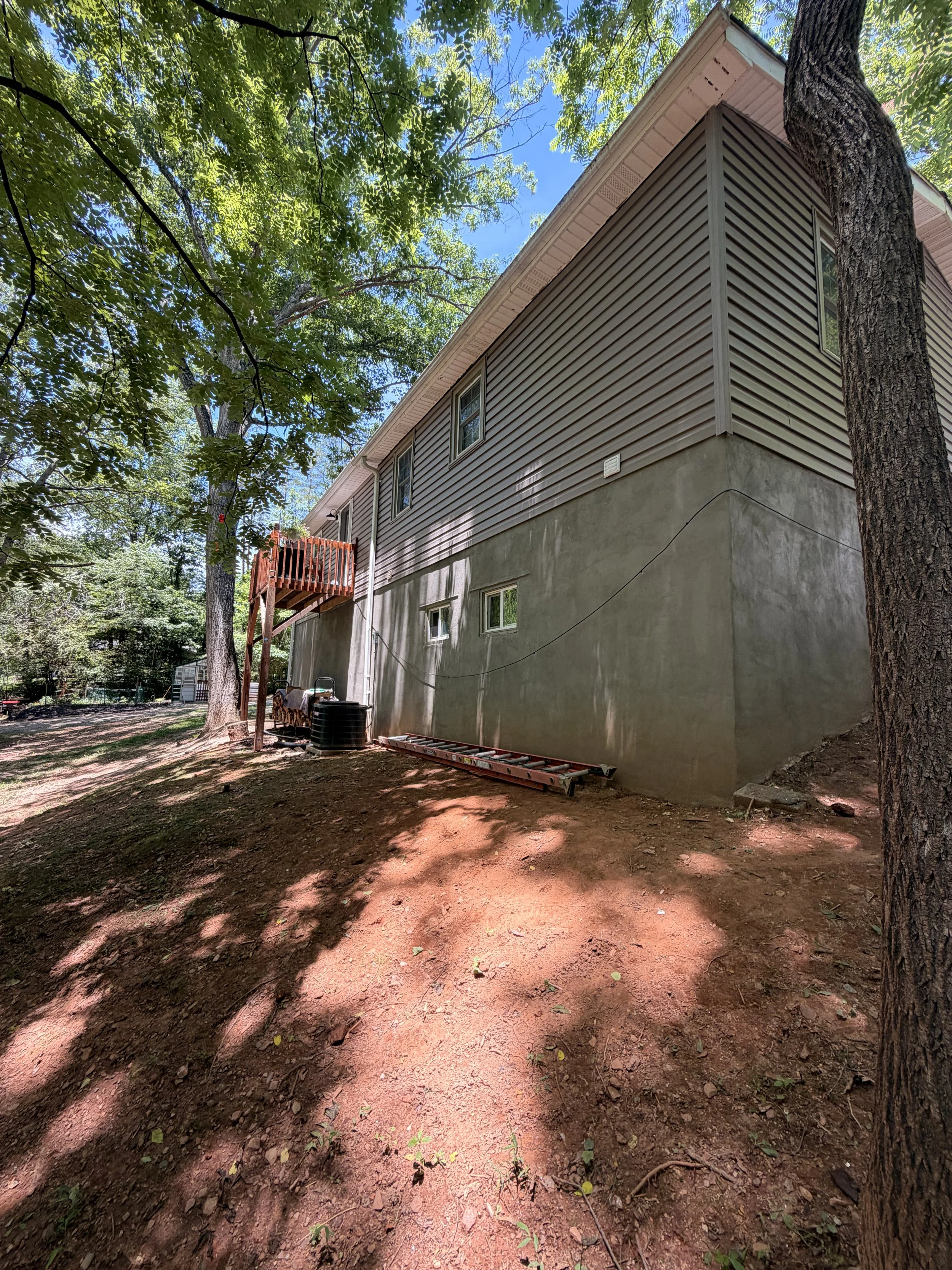 Backyard with dirt ground, large trees providing shade, a house with beige siding and a small deck, and construction tools and supplies near the house.