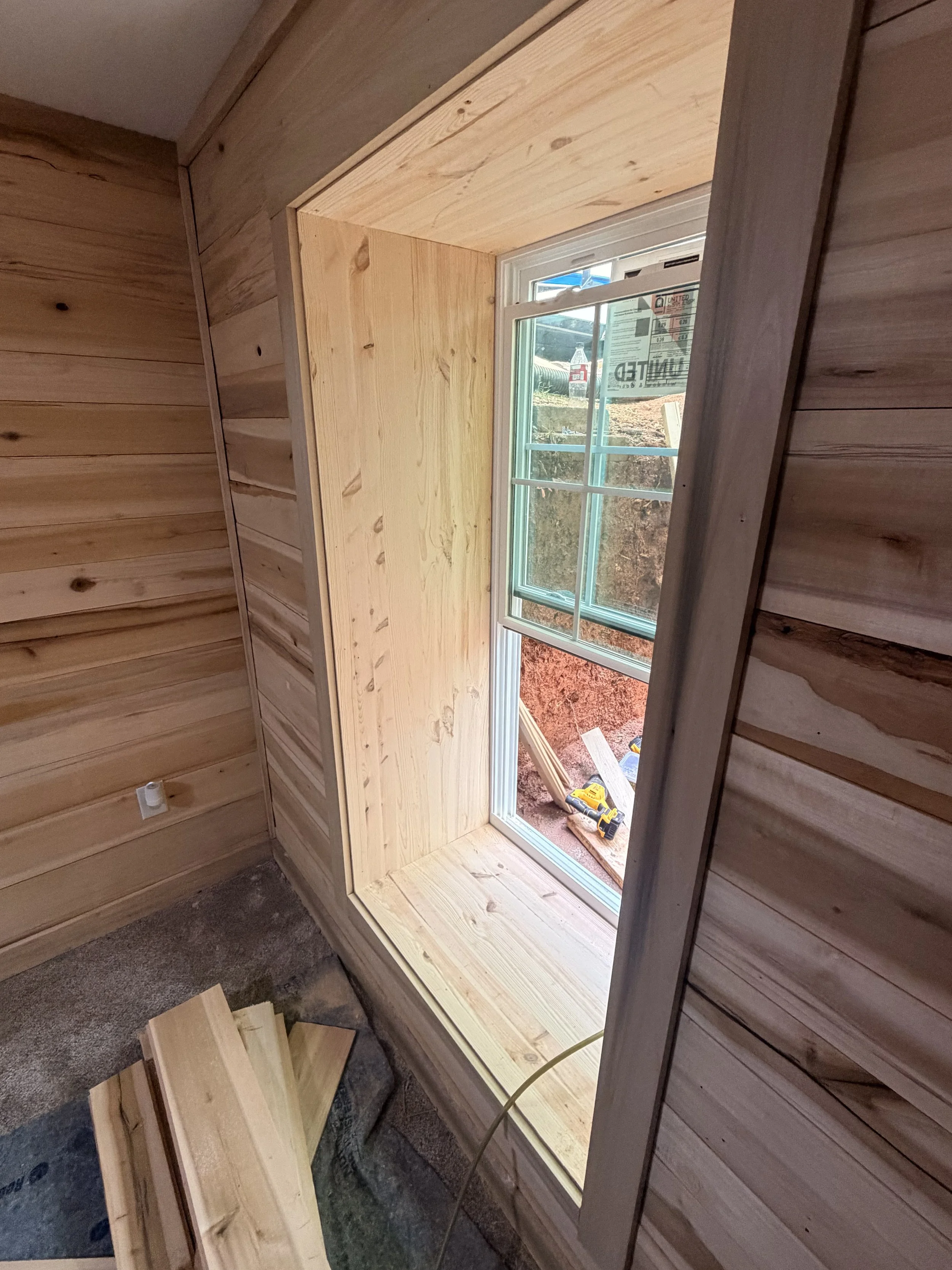 A window with a white frame set into a wooden wall under construction, with tools and construction materials on the ground outside.