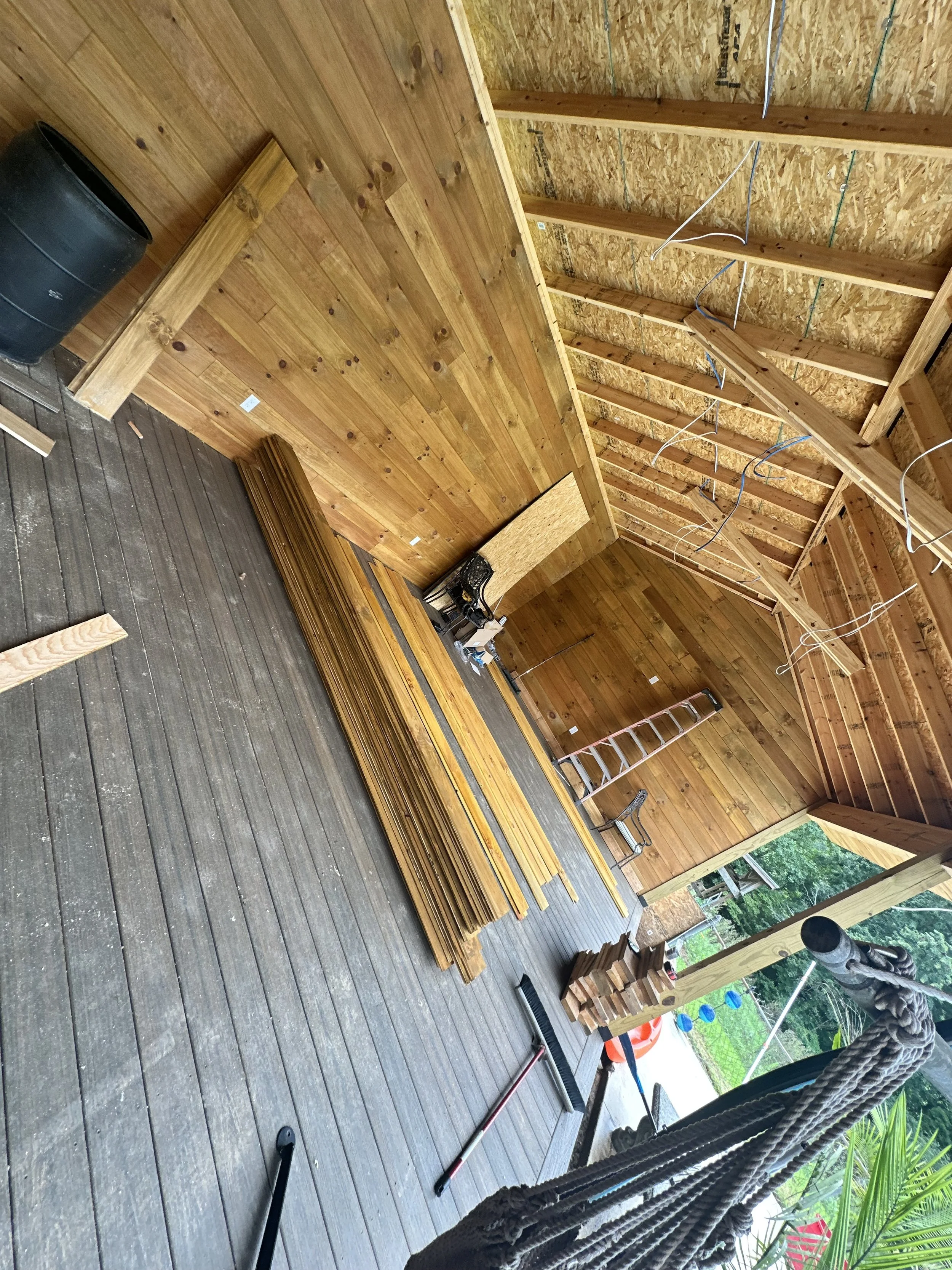 Photo of a construction site with wooden framing, a vertical wall made of planks, and building materials like wooden slats and tools on a gray porch floor. Several wires are hanging from the ceiling, and a black barrel is present near the wall. Outdoor greenery is visible in the background.