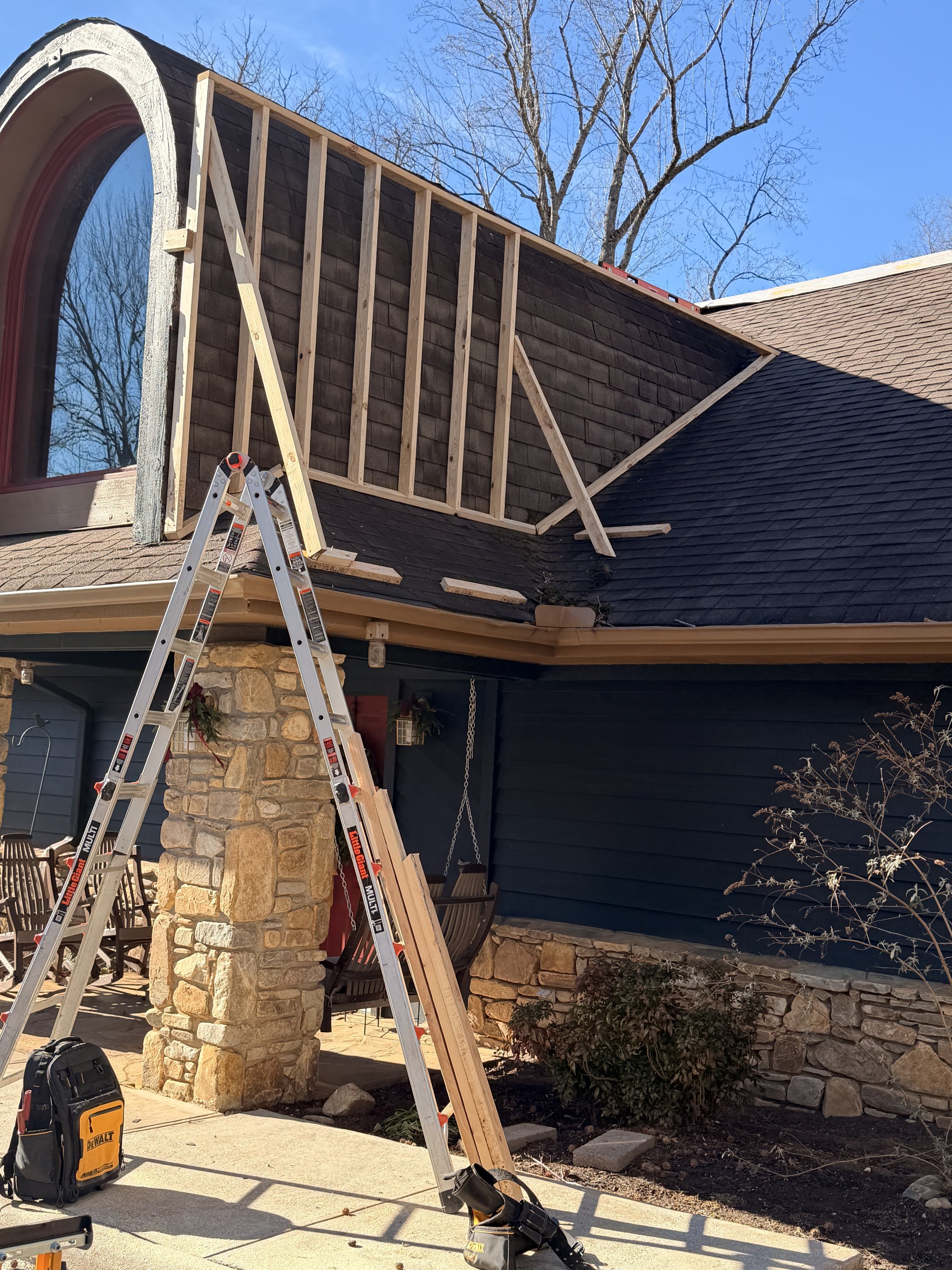 House exterior with a steep roof under construction, wooden beams, ladder, tools, and outdoor furniture near stone and dark siding wall.