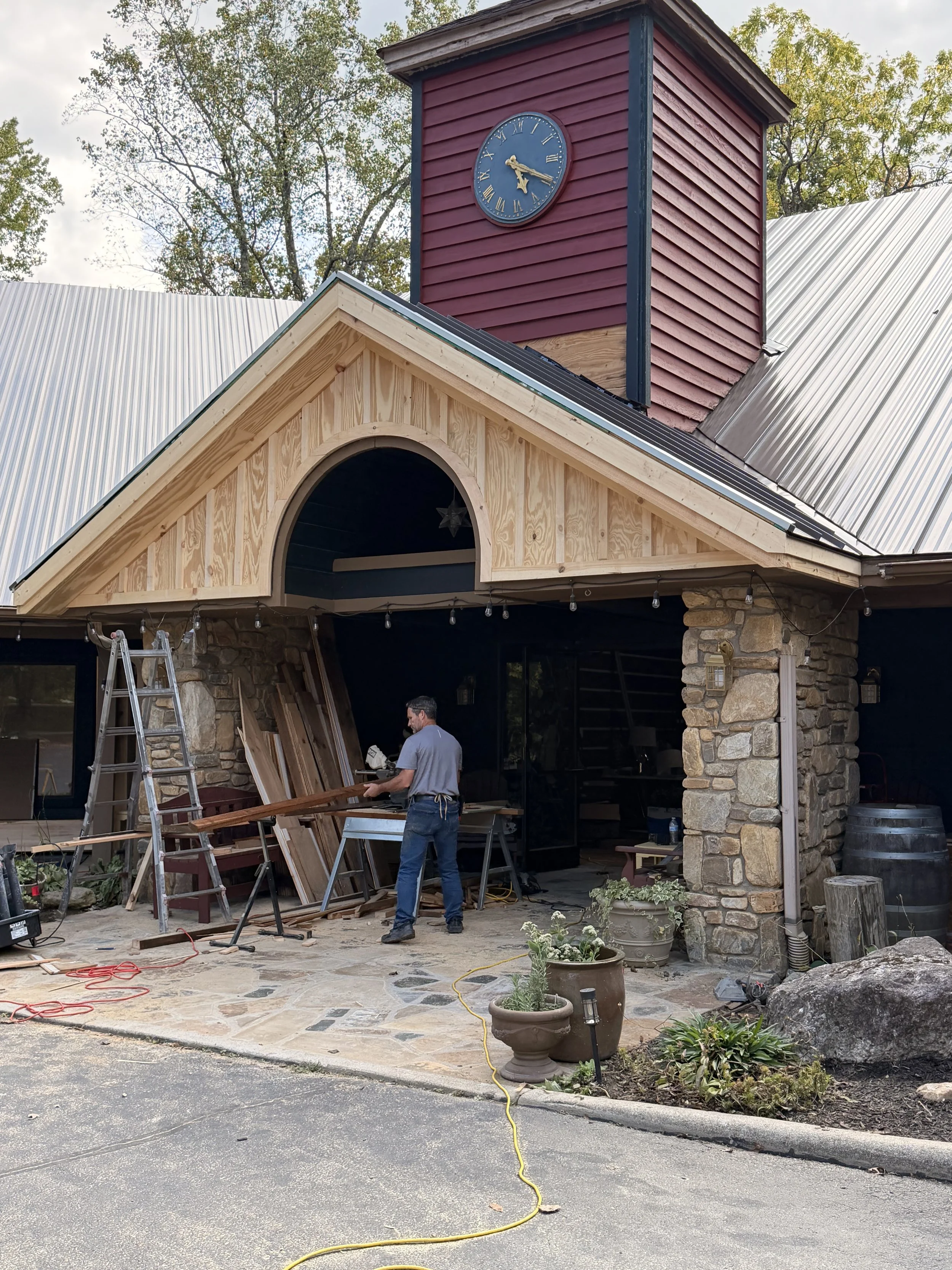 Construction workers installing a wooden beam on a building with stone and wood exterior walls. The building has a metal roof and a red clock tower. There are plants, a ladder, and construction tools and materials around.