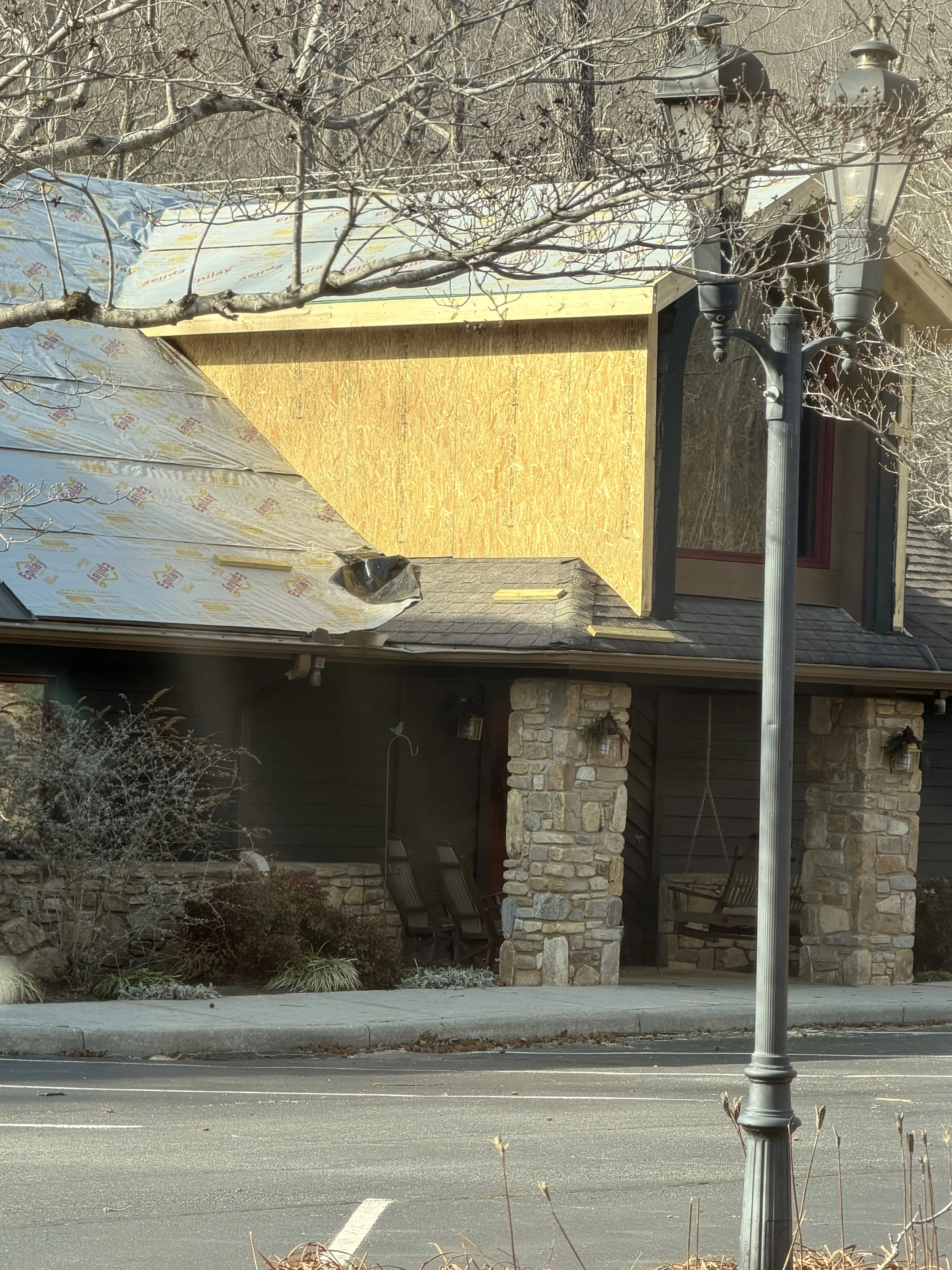 Front of a house under construction or renovation with plywood siding, brick pillars, a stone wall, and outdoor seating area with chairs. A streetlamp is in the foreground.