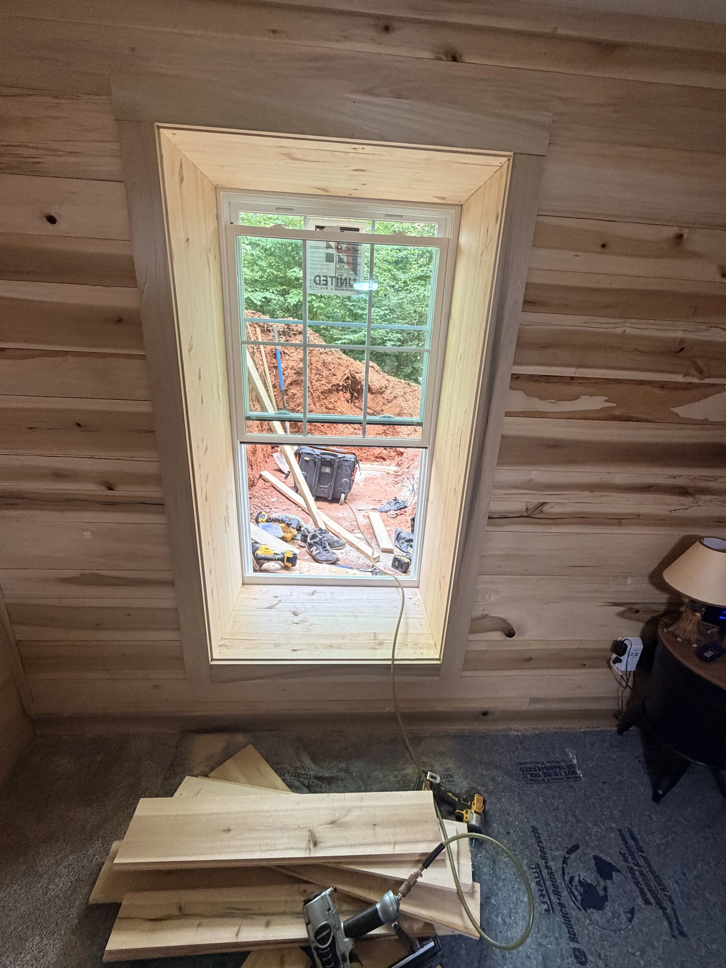 A construction site inside a room with wooden walls and a window, where woodworking and installation work is ongoing. Tools and materials are scattered around, including a drill, saw, and wood pieces.