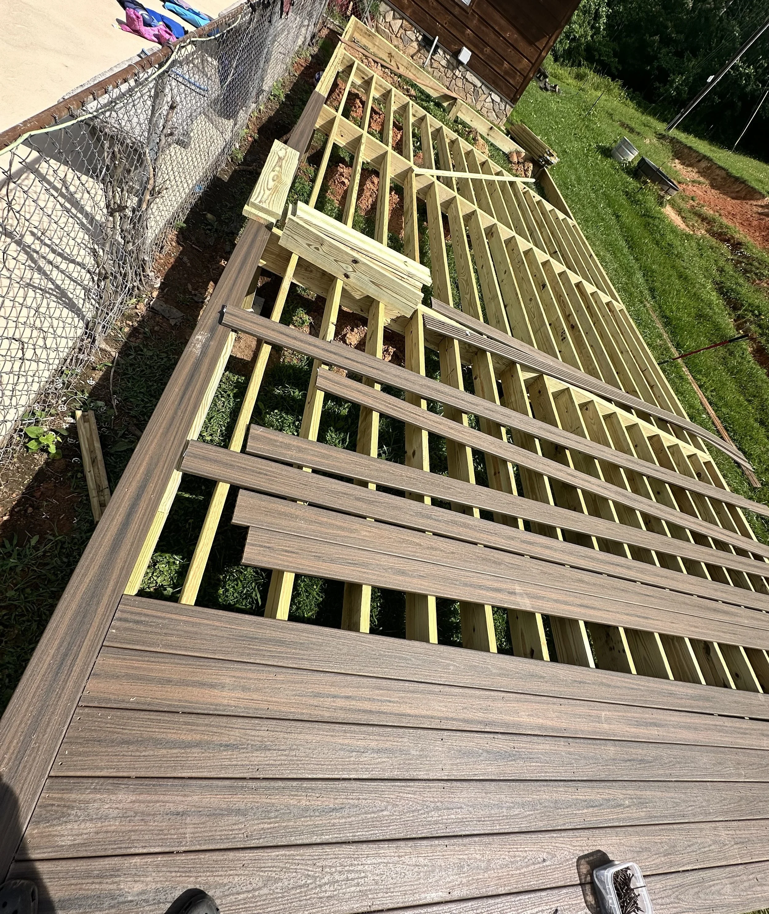 A wooden deck constructed with a framework of parallel and crosswise wooden beams, with some planks laid down. Construction tools and materials are visible around the deck, and a wire fence with hanging bags is along one side. In the background, there is a grassy area with potted plants and a small shed.