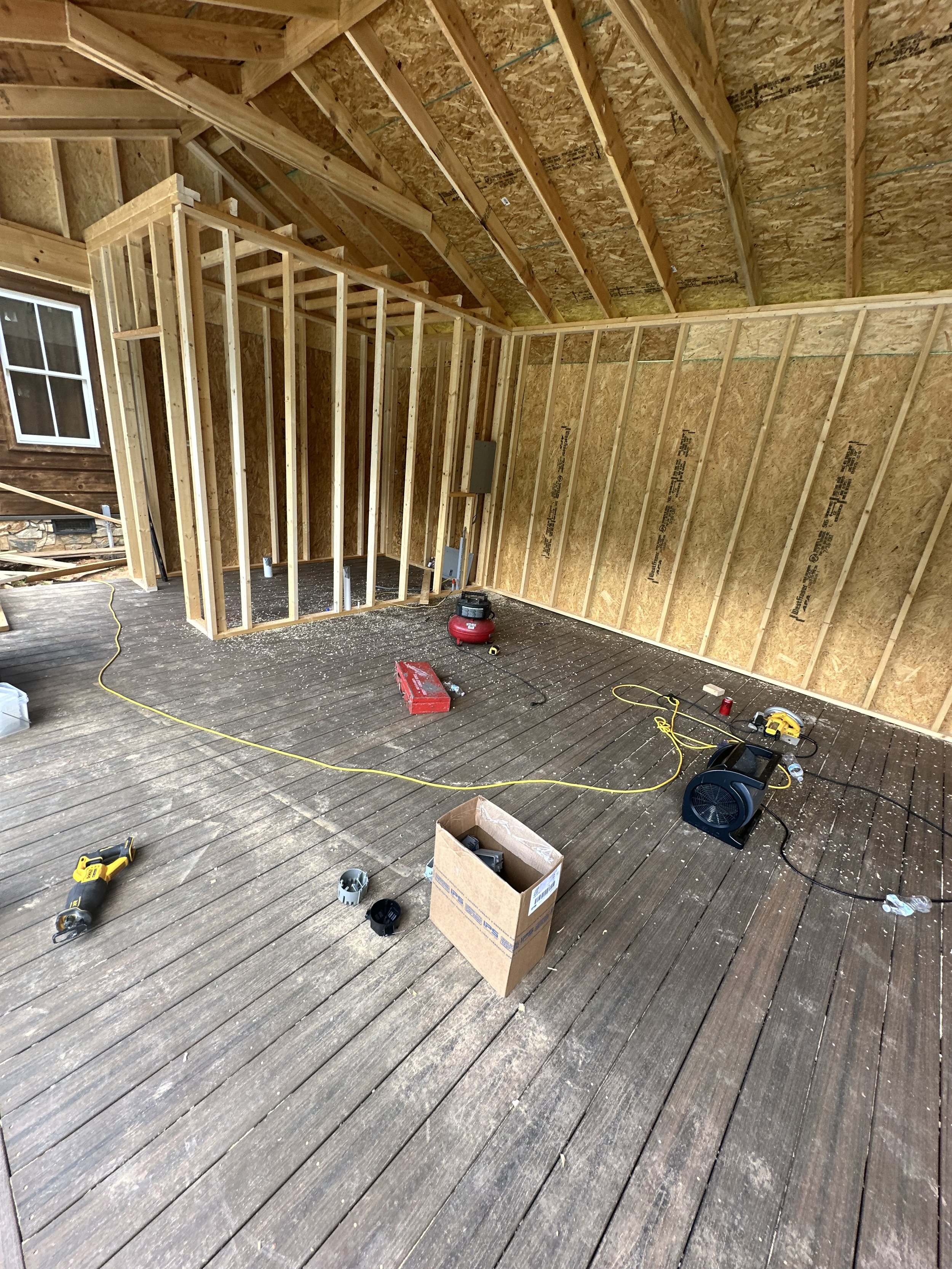 Construction site of a building with exposed wooden framing, an unfinished wall, and a ceiling. The floor is made of wooden planks with construction tools and materials scattered around, including a yellow drill, a red toolbox, a black fan, and several boxes.