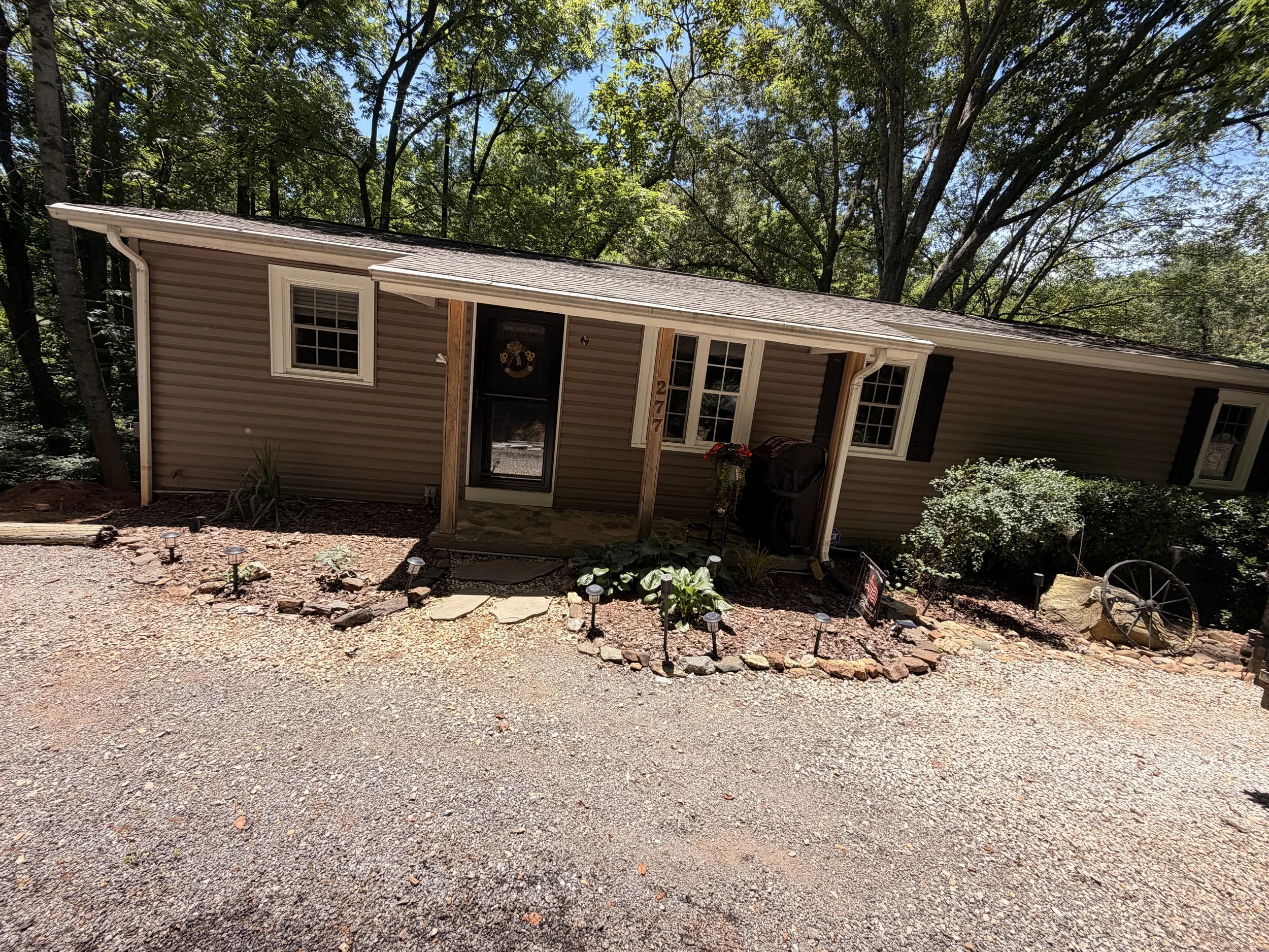 Photo of a small house with beige siding, white trim, a black front door with a decorative wreath, and a small porch with wooden posts. There are several windows, some with black shutters. The house is surrounded by a yard with gravel, small plants, and decorative lighting, with tall trees in the background and a blue sky.