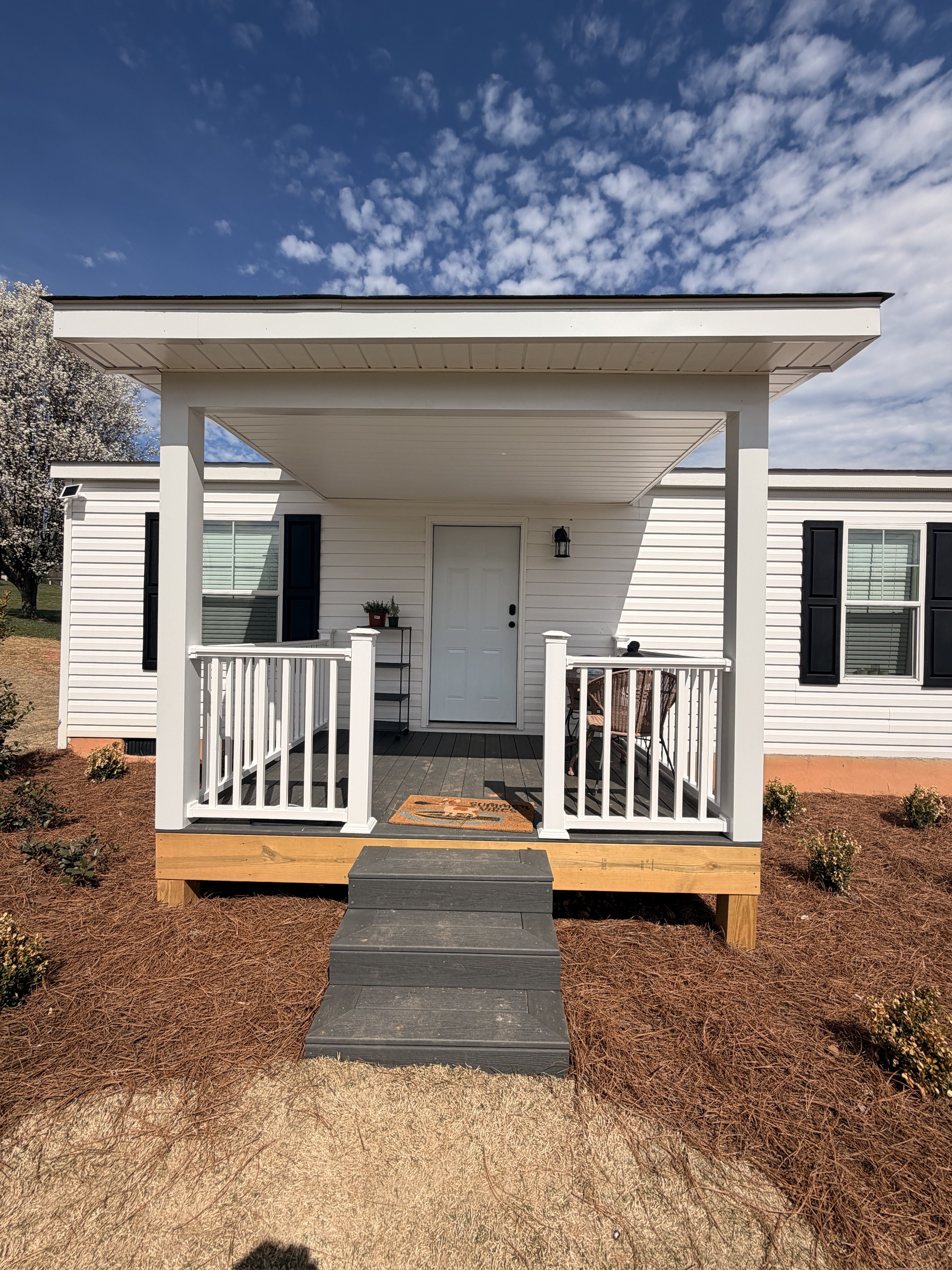 Front porch of a white house with black shutters, a small porch, steps, and outdoor furniture, under a blue sky with clouds.