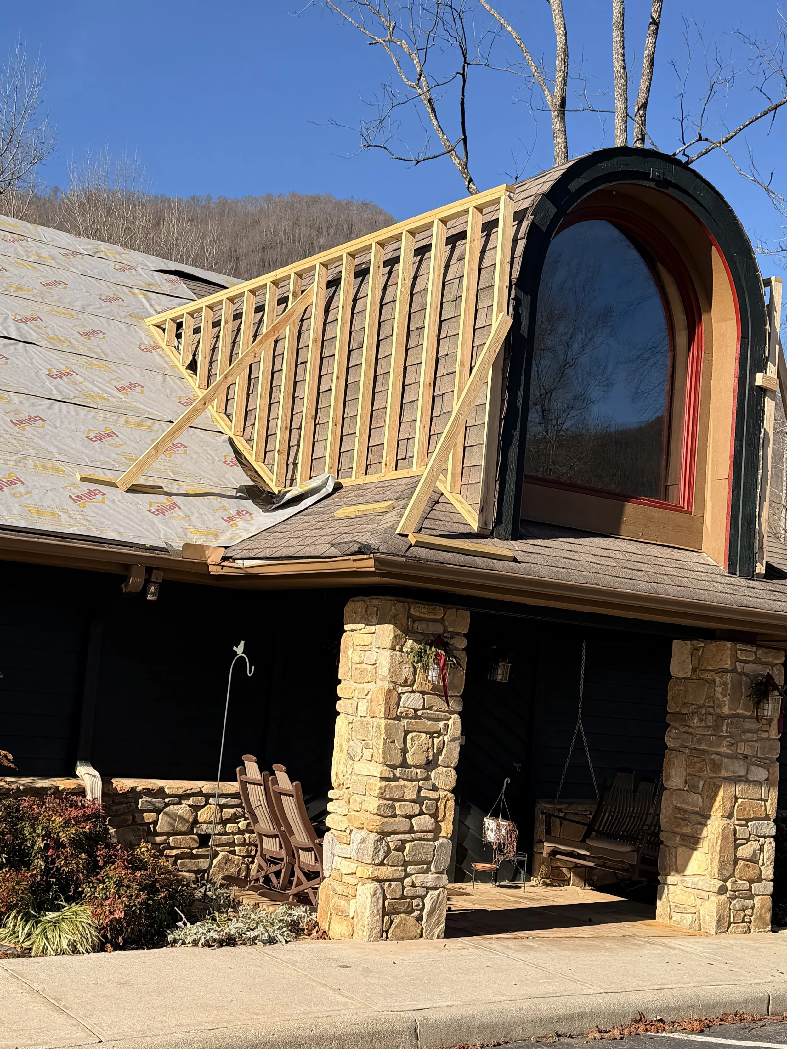A house under construction showing a roof with a dormer window and scaffolding, with stone pillars supporting a covered porch. There are outdoor chairs and plants in the patio area.