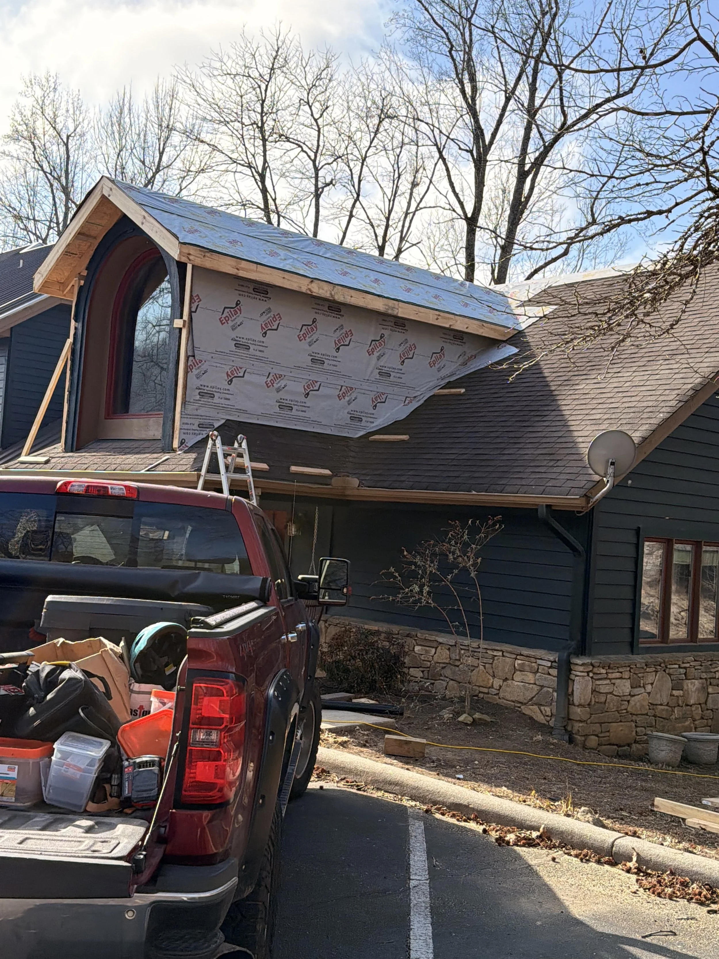 Construction work on a house with blue siding and a stone foundation. The roof is being replaced, with roofing materials visible. A ladder is leaning against the house, and a truck is parked in front filled with construction tools and supplies.