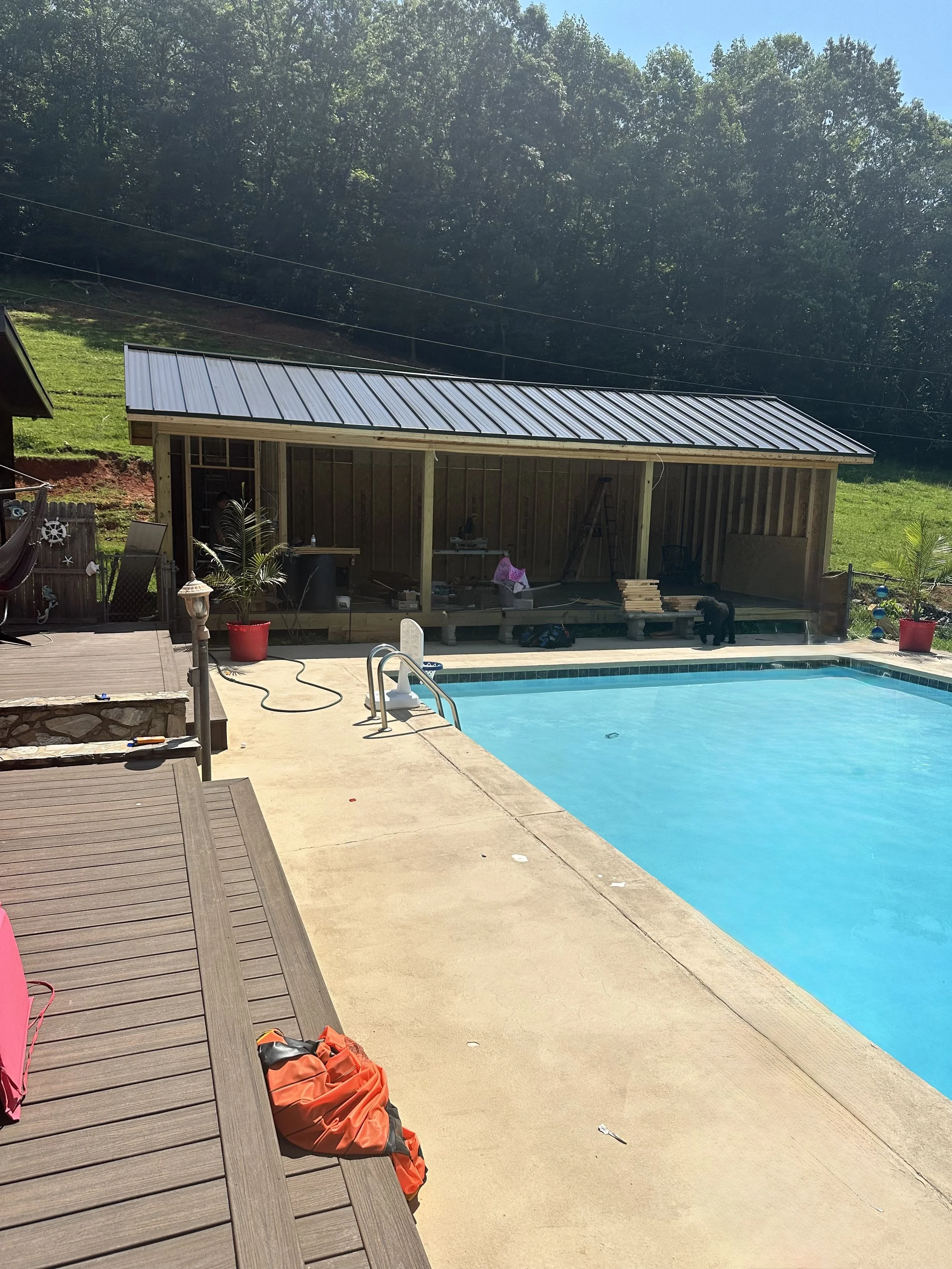 A backyard scene with a swimming pool, a wooden deck, and a partially constructed wooden shed with a metal roof. There are some tools and construction materials around, along with a black dog near the shed. The background features a grassy hill and a wooded area under a clear blue sky.