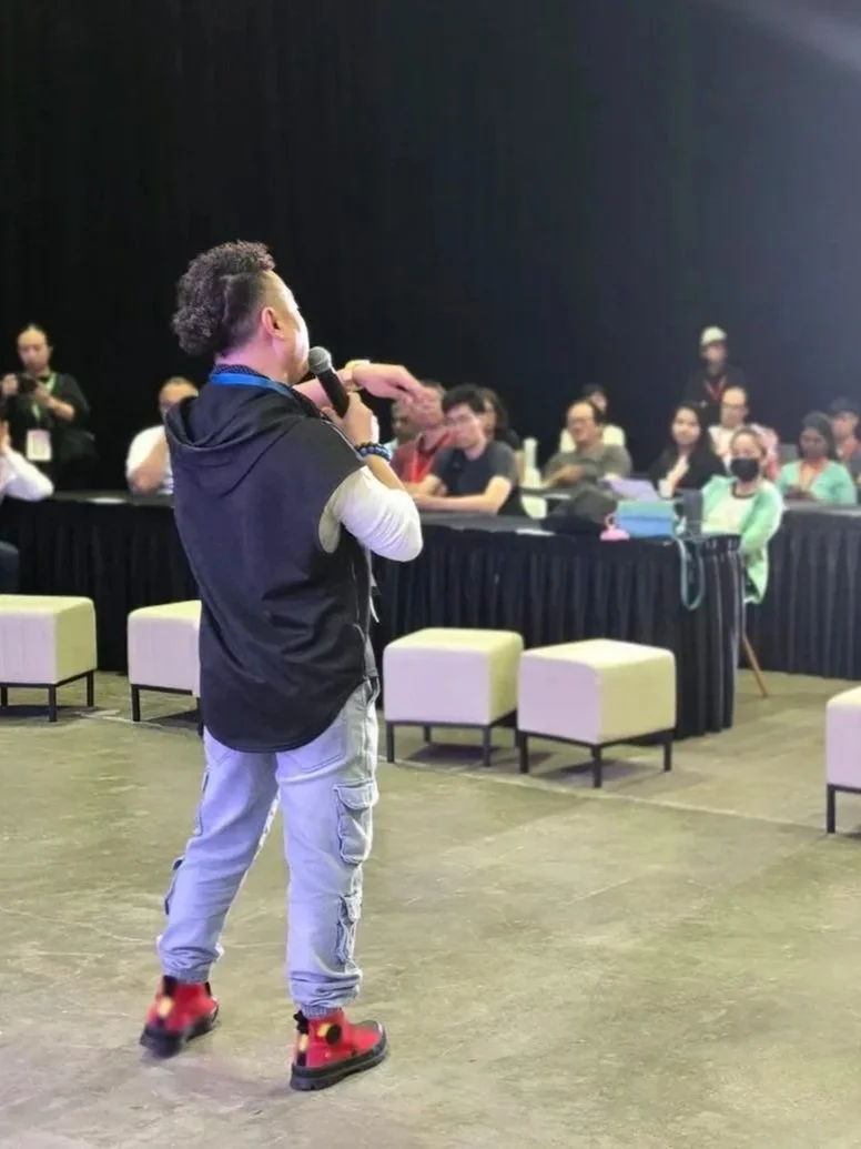 A man is speaking into a microphone in front of a seated audience at an indoor event. The audience members are listening, some wearing masks, with a black backdrop behind them. The man has short curly hair and is dressed casually in a black vest, white long-sleeve shirt, light cargo pants, and red sneakers.