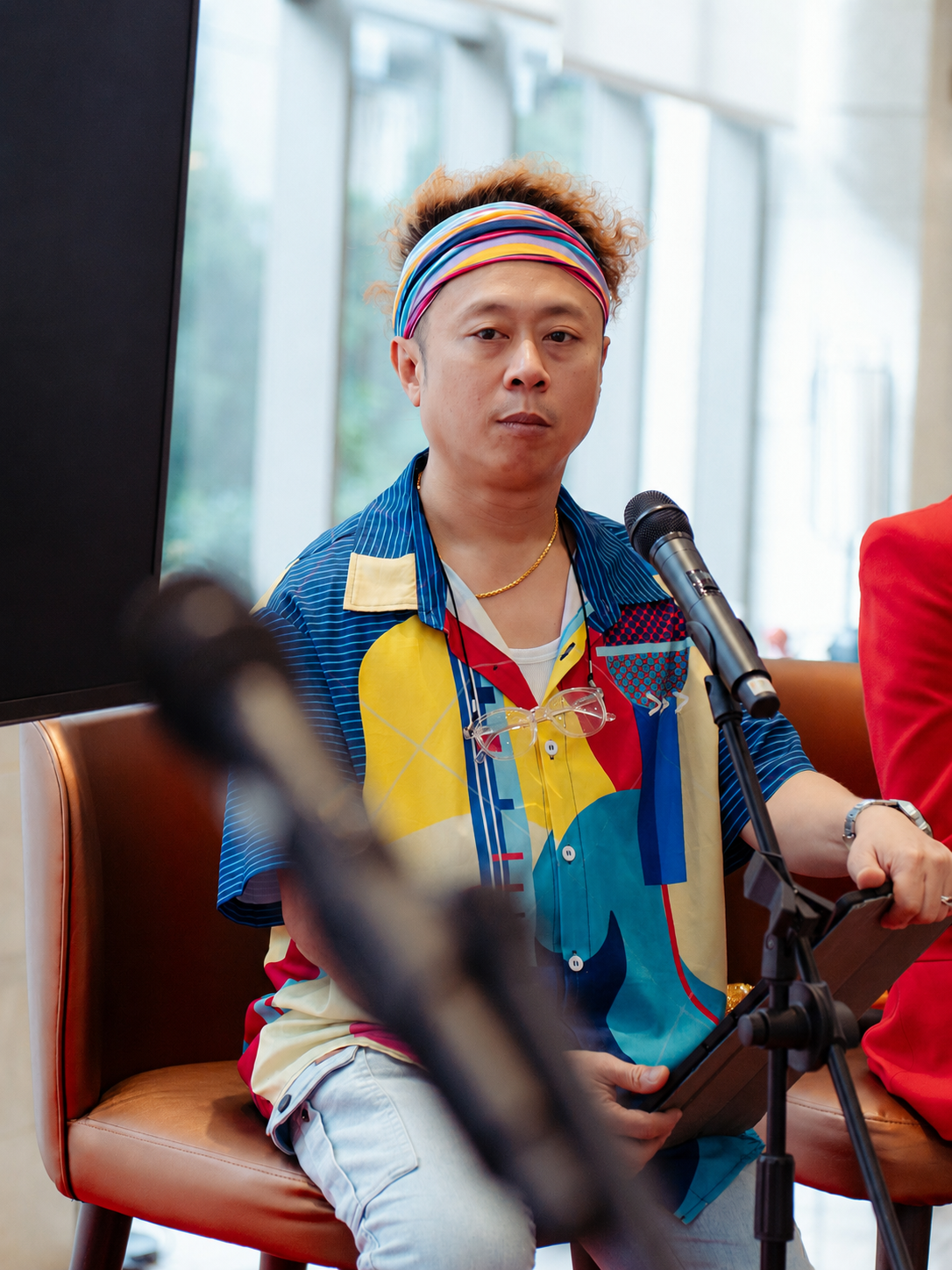 Jason Elpheus Wong, wearing a colorful headband and a vibrant, patterned shirt, sitting at a table with a microphone in front of him, holding a tablet or book, in an indoor setting with large windows in the background.