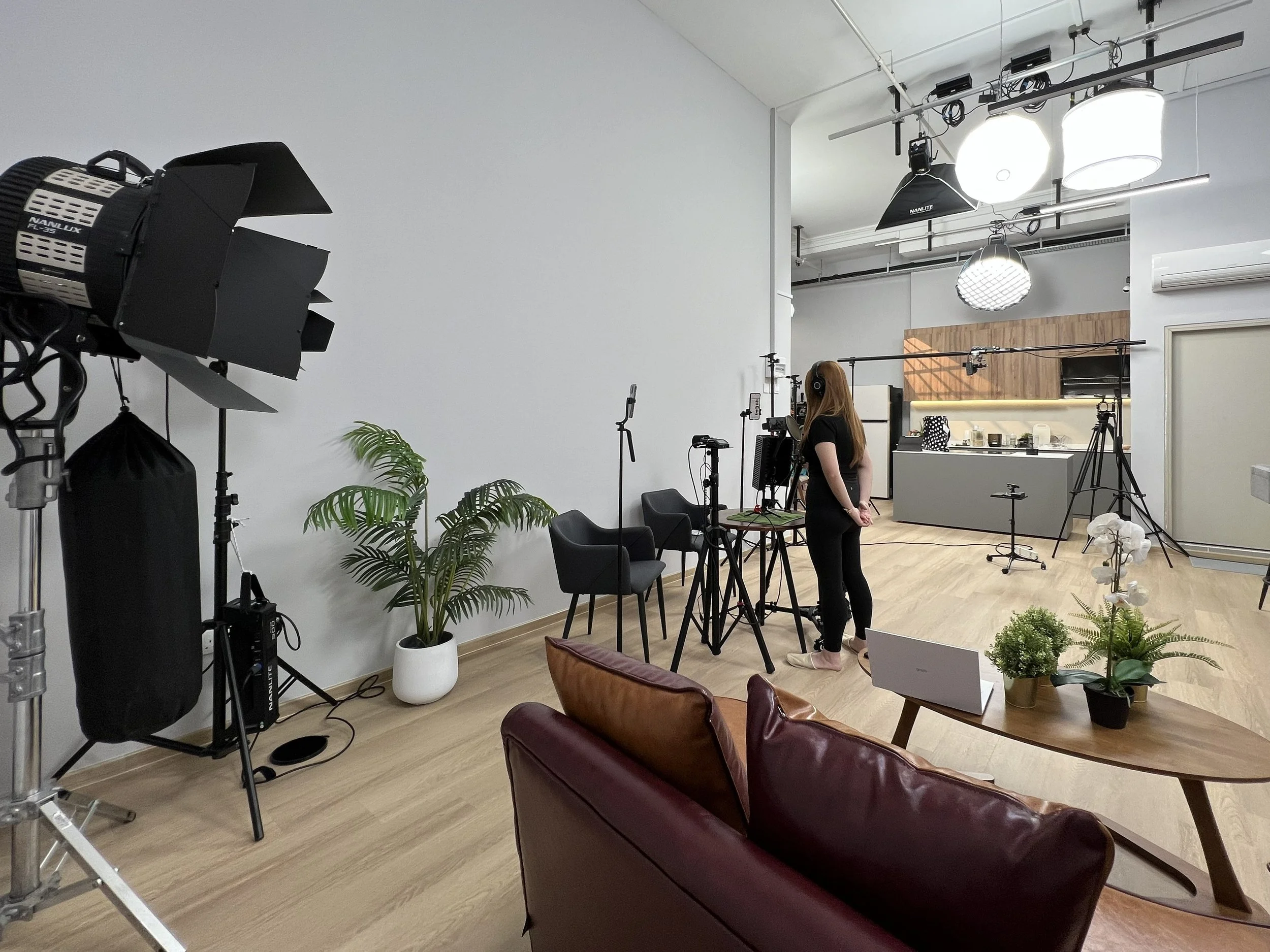 A person wearing headphones standing in a photography or video studio with professional lighting and camera equipment. The studio has plants and seating area with chairs and a brown leather sofa.