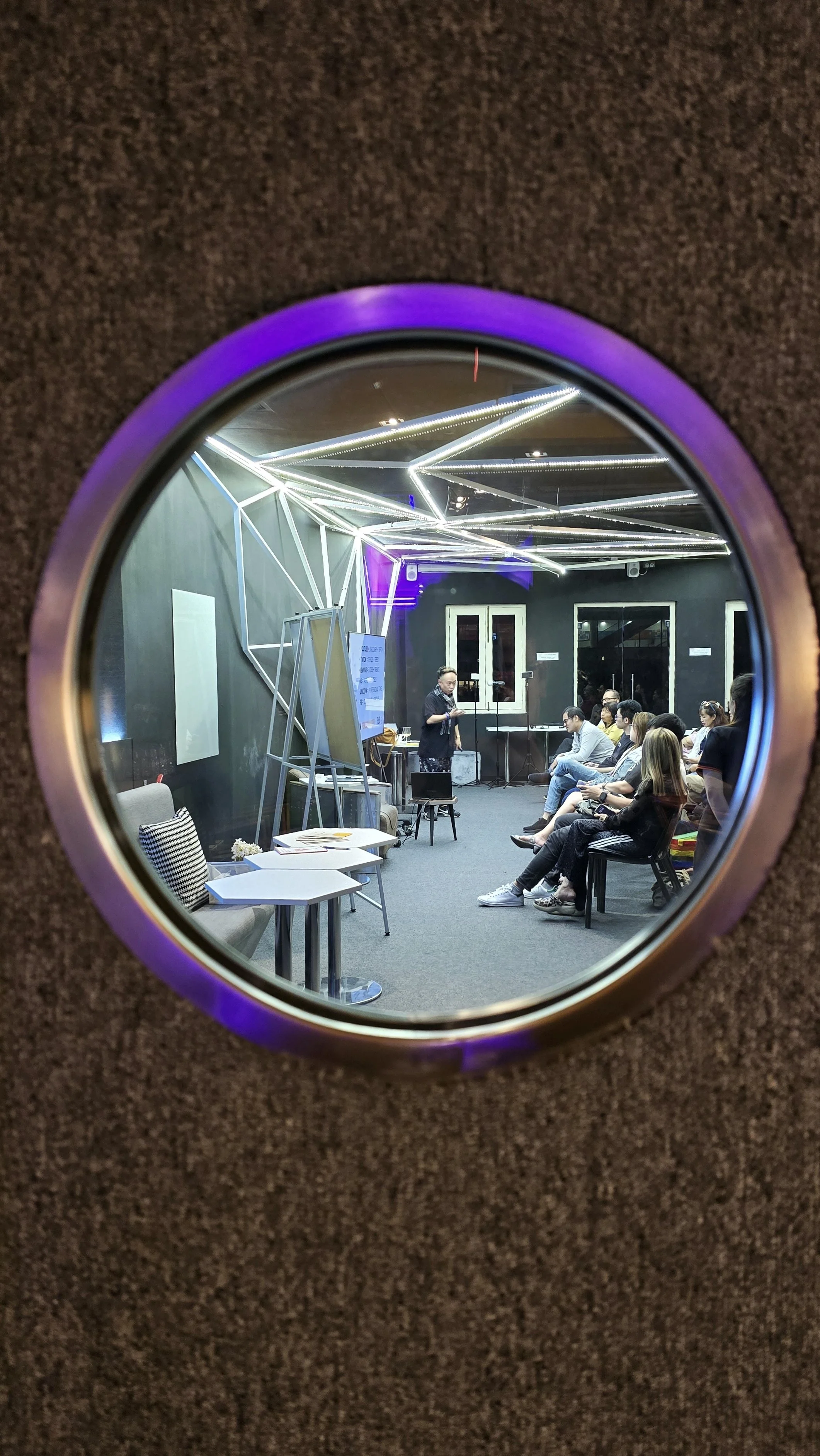 Audience watching a speaker in a modern, neon-lit room, viewed through a porthole-style window.