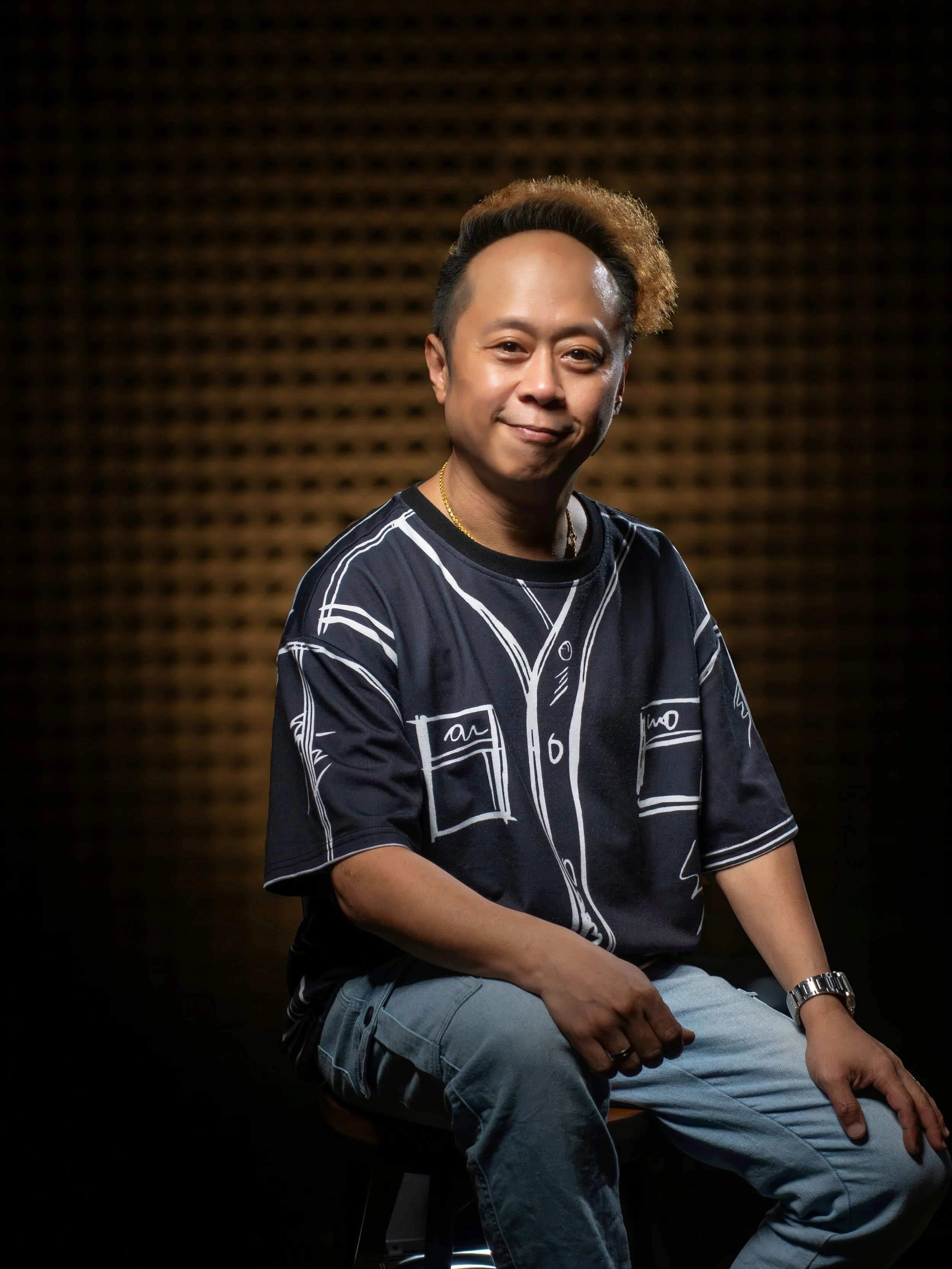 Portrait of a Jason Elpheus Wong sitting on a stool against a dark, textured background, wearing a navy blue shirt with white line drawings, light blue jeans, a watch, and rings, smiling with a relaxed expression.