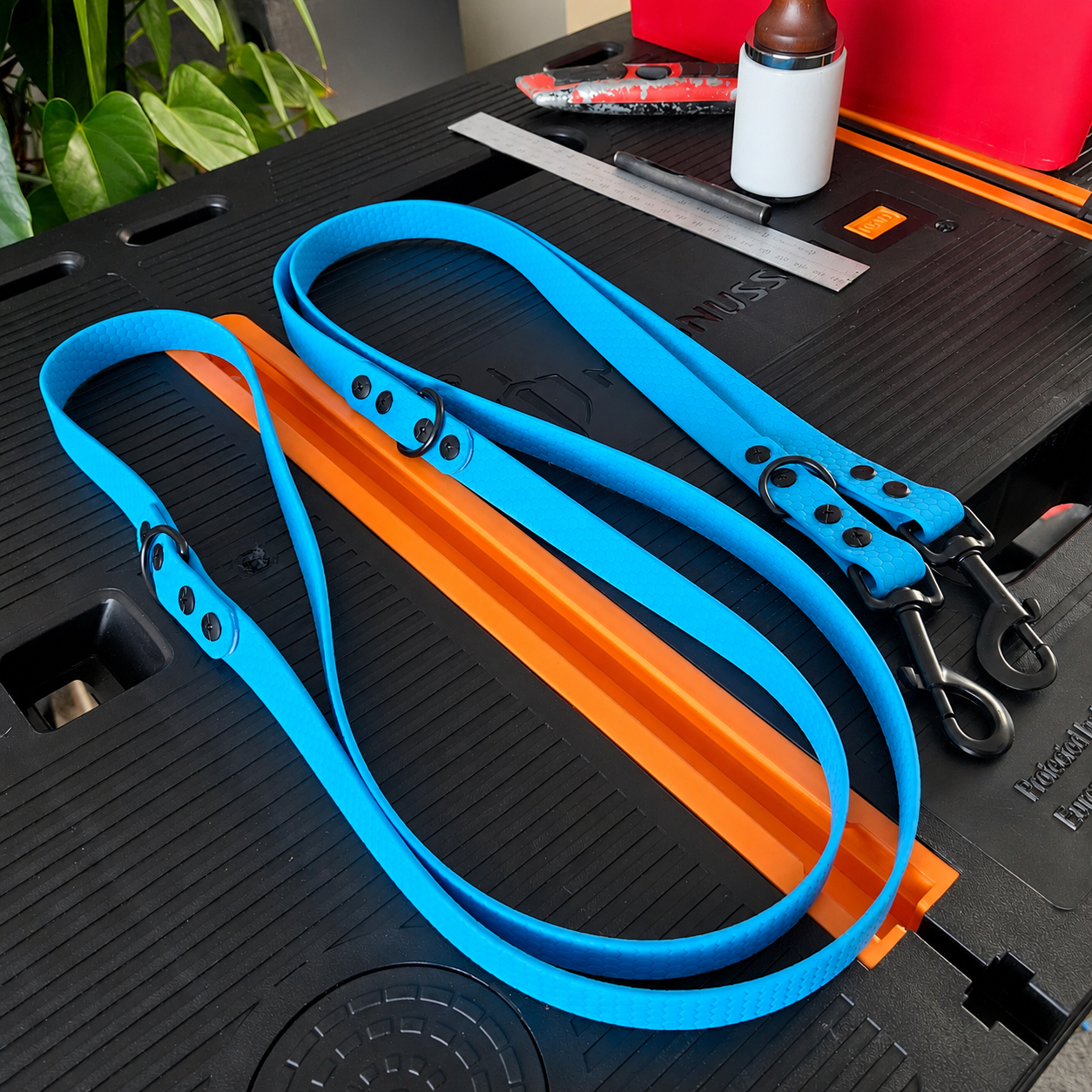 Two blue pet leashes with metal clips on a black workbench with an orange guide rail, a metal ruler, a black marker, a white bottle with a brown cap, and a red container in the background.