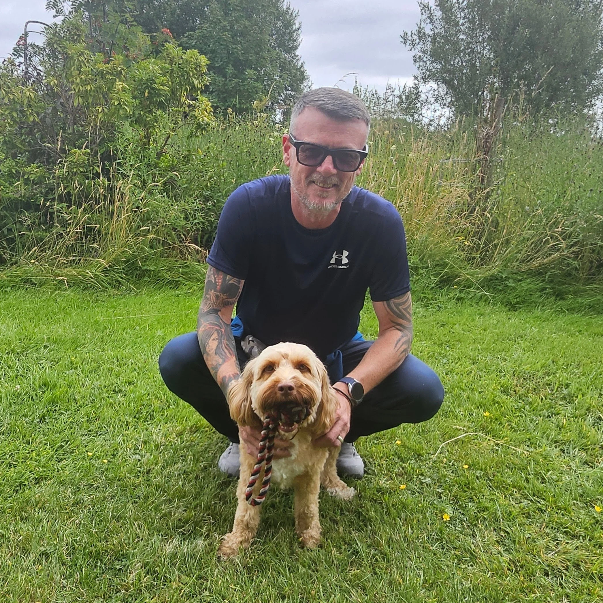 A man with tattoos, sunglasses, and a beard, crouching on grass in a lush outdoor setting with greenery and trees, holding a brown and white dog on a leash.