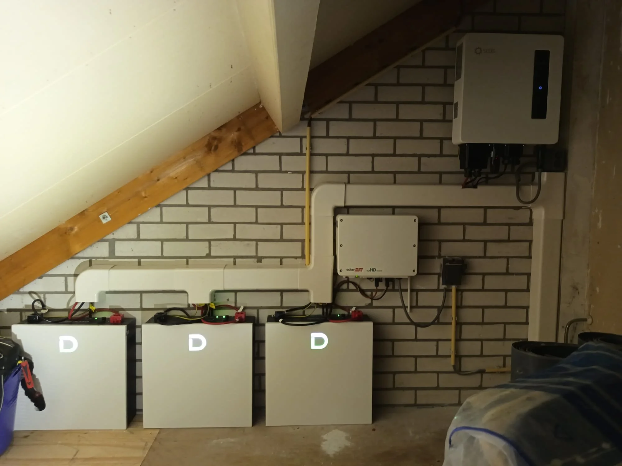 Elektrisch installatie met drie omvormers, aangesloten op een muur met bakstenen, onder een schuine plafond. Drie stuks LV batterijen van het merk Dyness met een hybride omvormer van het merk Solis.